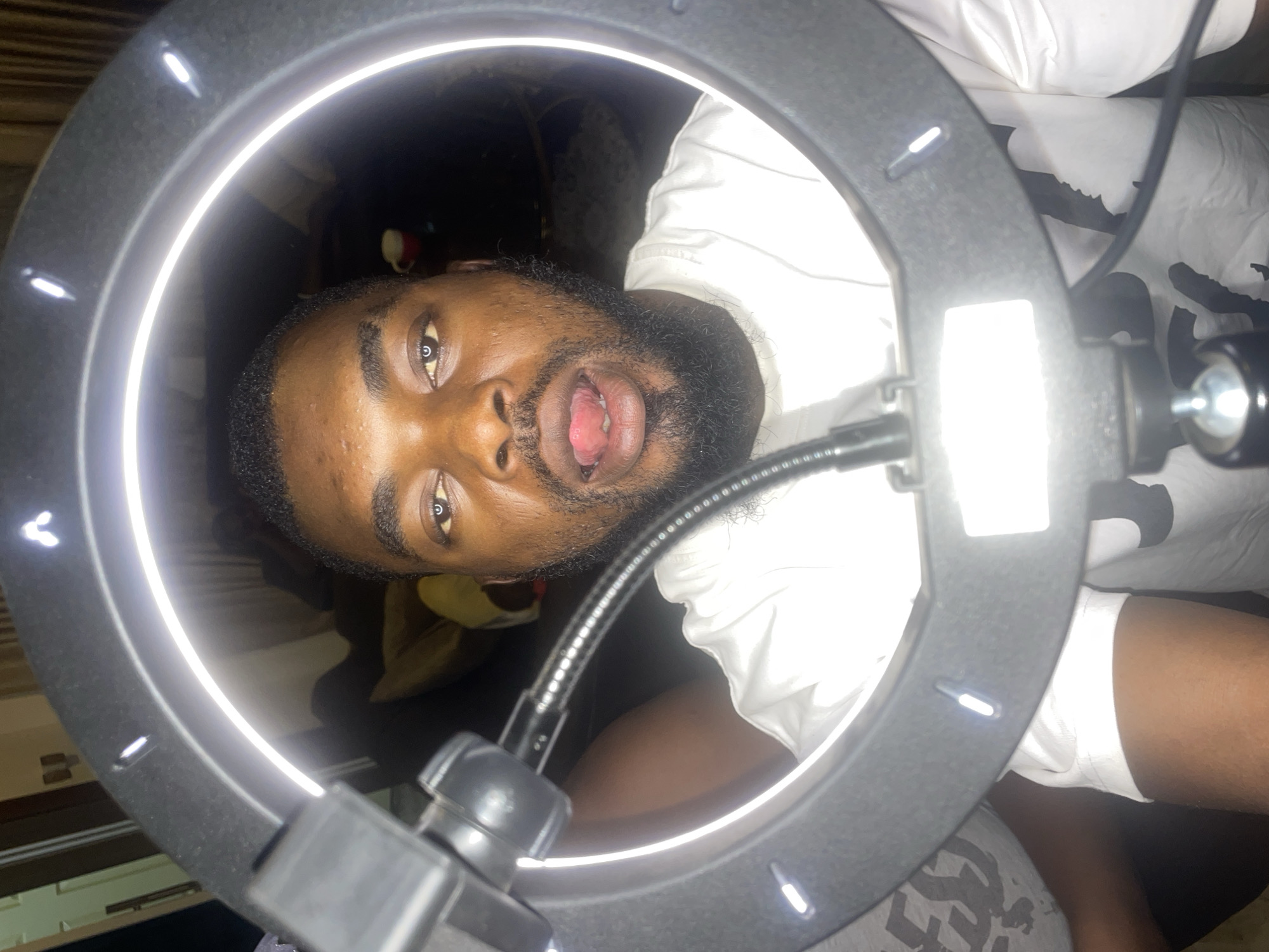 A man with brown skin, dark hair, and a beard is centered in the frame. He is positioned within a ring light. He is wearing a white t-shirt. His mouth is open. The background behind him is dark and out of focus. The photo was taken in Jos, Nigeria.