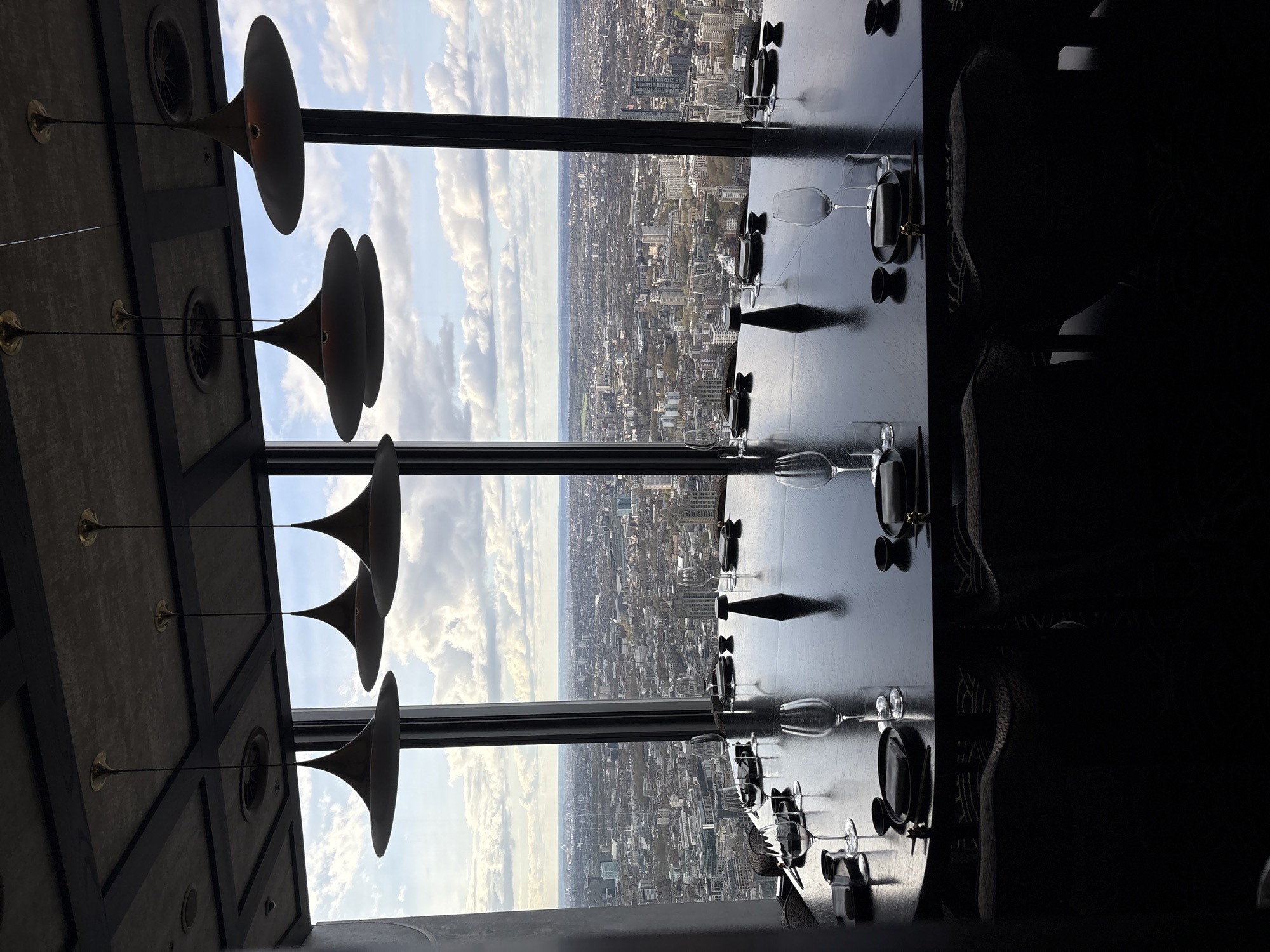 The image shows a table setting in a restaurant high above the city of London, United Kingdom. Outside the window, the London skyline is visible, with a mix of buildings and green spaces under a cloudy sky. The table is set with wine glasses, plates, and cutlery. The lighting fixture hanging from the ceiling is visible on the left. The right side of the image is dark, showing seating.