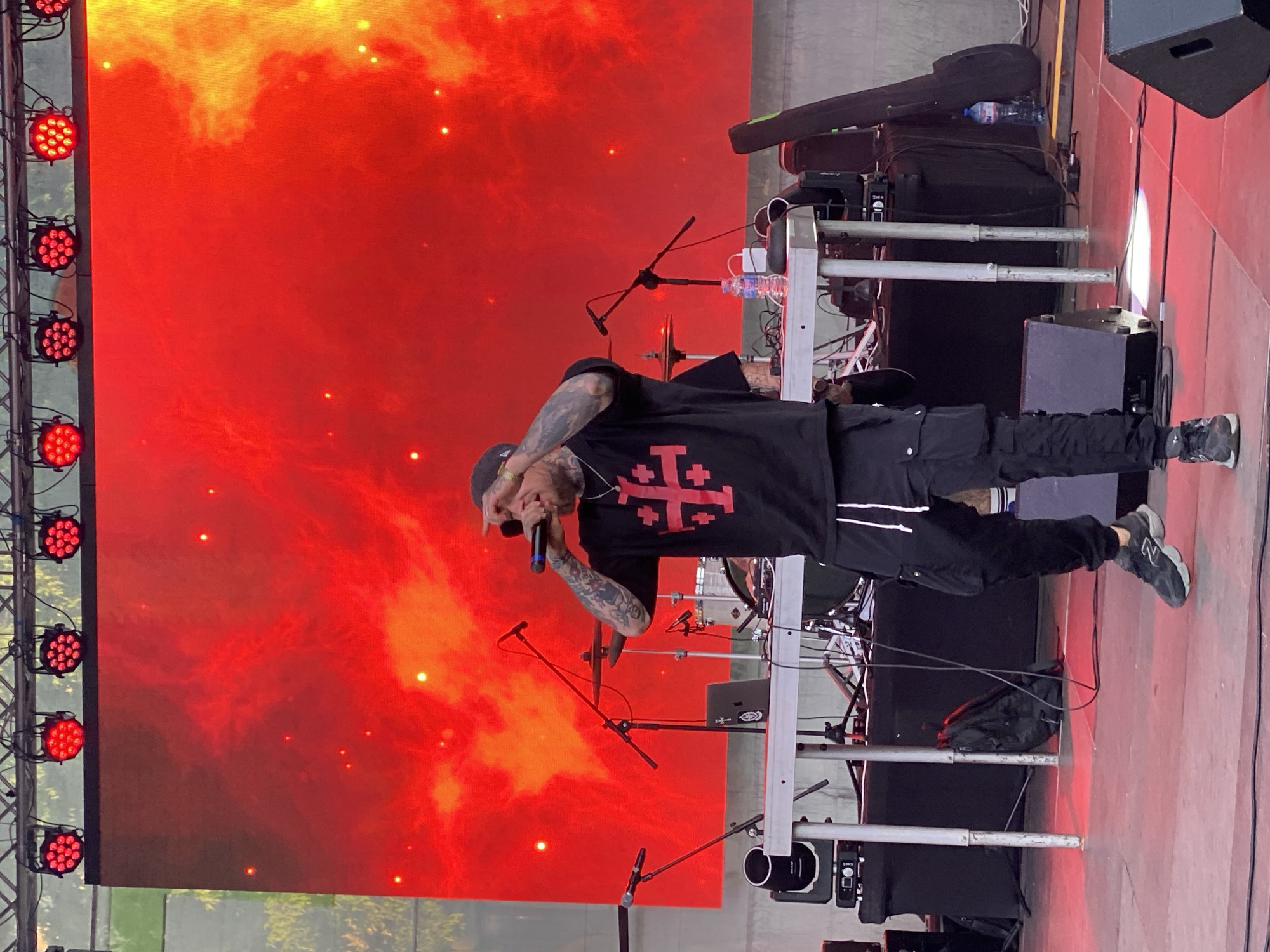 A singer stands on a stage illuminated by a red backdrop and red stage lights. The stage appears to be outdoors, possibly in a park, as some greenery can be seen at the bottom of the red screen. The singer, wearing a black t-shirt with a cross design, black pants with white stripes, and sneakers, is holding a microphone in his right hand and appears to be performing. The stage is equipped with musical instruments, including what looks like a keyboard setup.