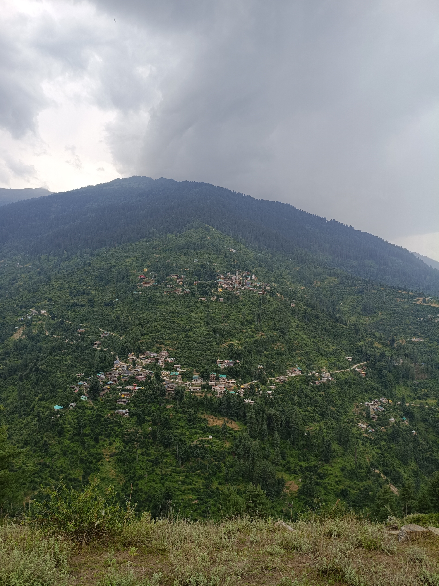 The image captures a high-angle view of a village nestled on a steep, densely forested mountainside in Chhaki, India. The village appears to be comprised of numerous scattered houses with predominantly light-colored walls and red or green roofs. The houses are situated along winding paths and roads that navigate the challenging terrain. The mountainside is covered in a mix of coniferous and deciduous trees, with some terraced fields visible, indicating agricultural activity. The sky is overcast with heavy, dark clouds, suggesting impending rain or a stormy atmosphere. The foreground of the image shows dry, grassy vegetation and soil, suggesting a higher vantage point overlooking the village. There are no people visible in the image.
