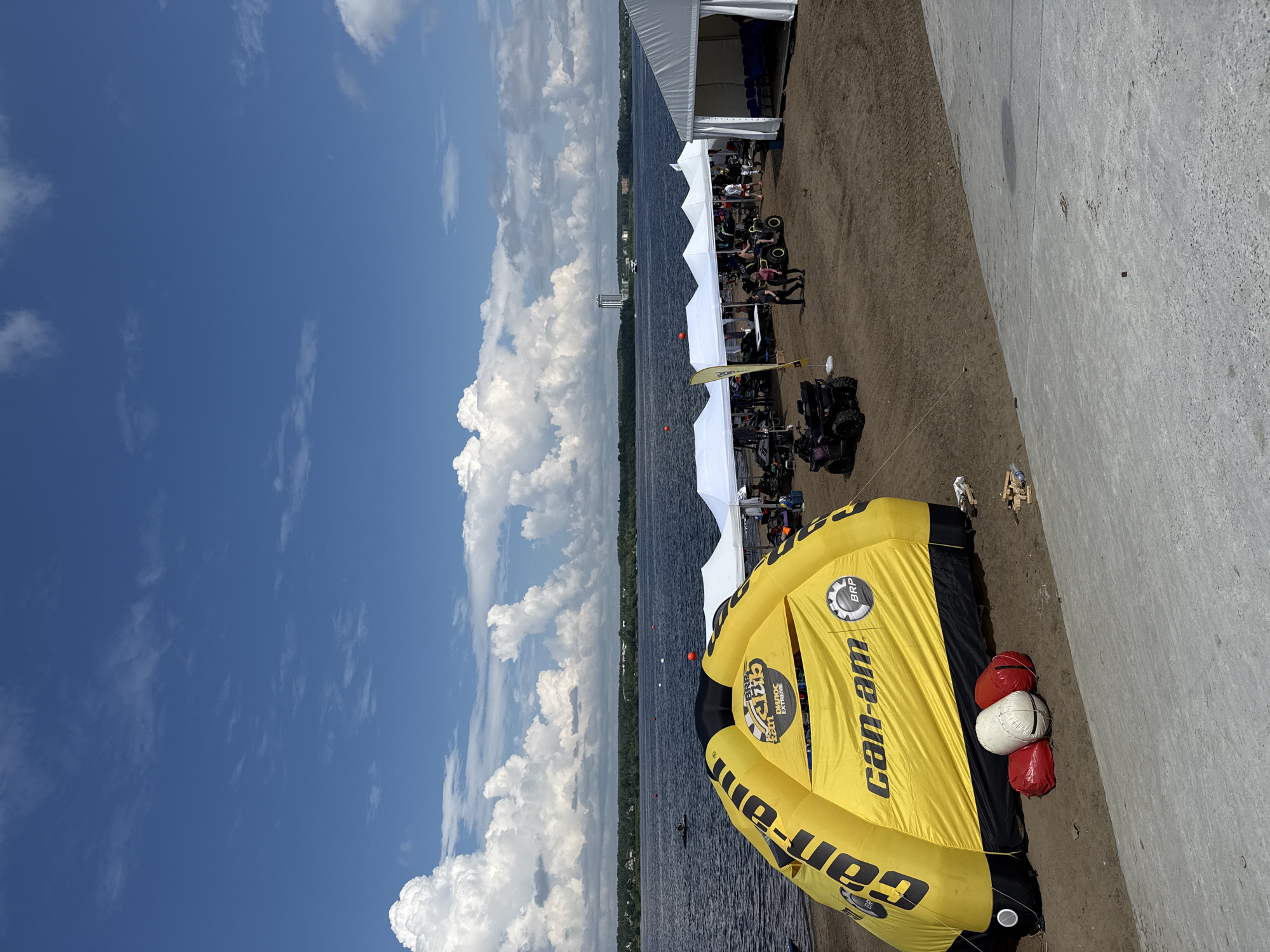 The image shows a waterfront scene in the city of Perm, Russia, on a bright, cloudy day. In the foreground, a large, inflatable yellow tent with "Can-Am" branding is set up on a sandy beach. Three large cylindrical bags, two red and one white, are placed in front of the tent. Beyond the tent, several white pop-up tents are arranged, with people milling around and ATVs parked nearby. The water of the Kama River stretches out to the horizon, with trees and buildings visible on the distant shore. Several small boats are on the water, and a tall building stands out against the skyline. The sky is a vibrant blue, dotted with large, fluffy white clouds. The overall scene suggests an outdoor event or festival taking place by the river.