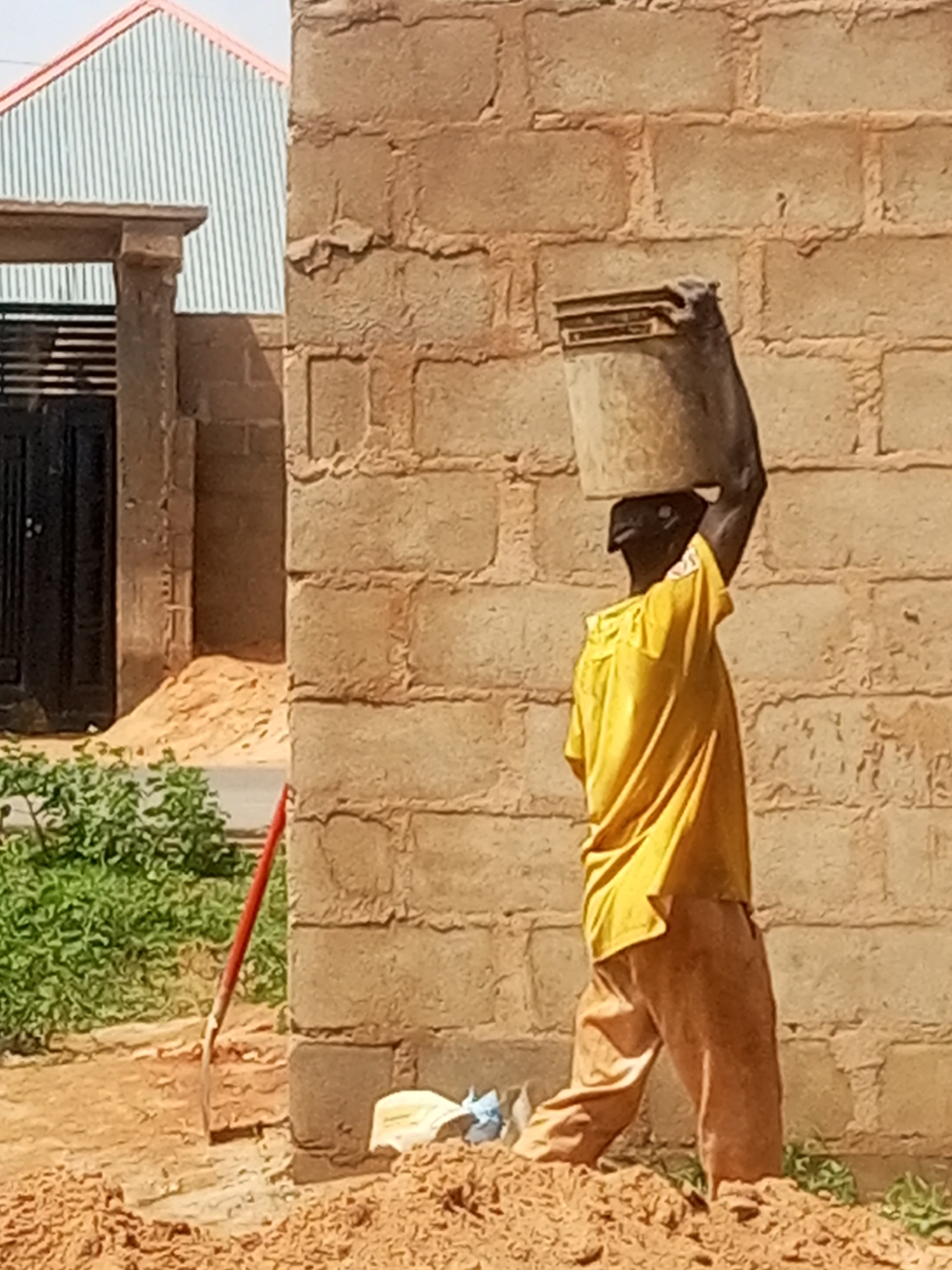 A person wearing a yellow shirt and brown pants carries a bucket of material on their head. The person is standing on loose dirt in front of a brick wall. To the left of the person, a shovel with a red handle leans against the wall. In the background, a building with a corrugated metal roof and a black door is visible. The environment appears to be an outdoor construction site.