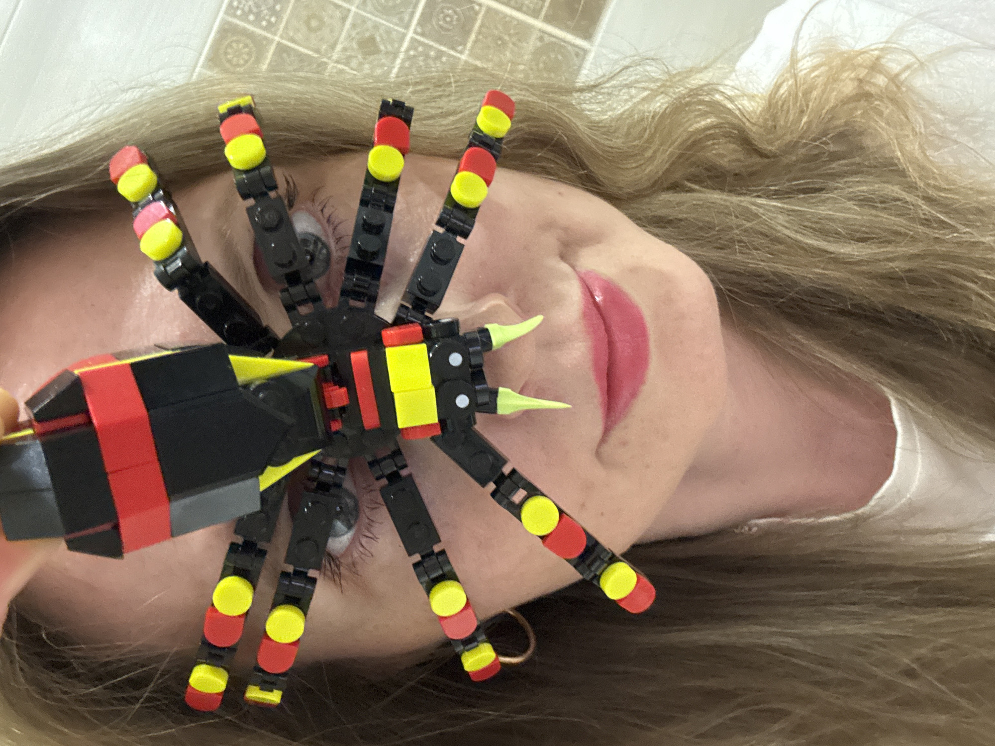 A person is lying down with their head tilted, and a Lego spider is positioned over their face. The spider is predominantly black with red and yellow accents on its legs and body. The person has blond hair, fair skin, and pink lipstick. The background appears to be a tiled surface. The location in the city of Chaikovsky, Russia is unknown.