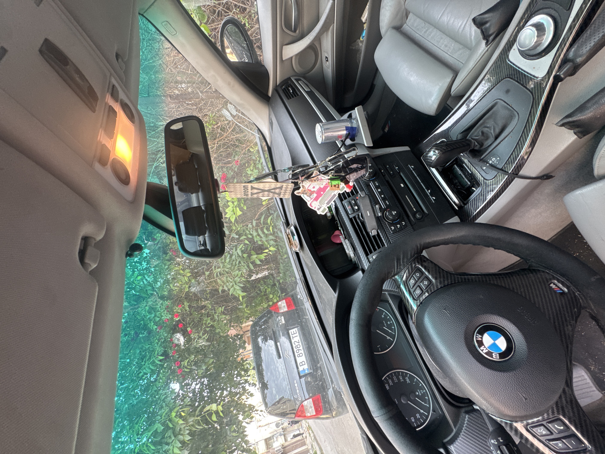The media file captures the interior of a BMW car, specifically from the driver's perspective looking through the windshield. The dominant feature in the foreground is the black leather steering wheel, adorned with a BMW logo at its center and an "M" logo, along with carbon fiber accents and various control buttons. The instrument cluster shows numerical markings such as "20", "40", "180", "200", "220", "240", and "260".
The dashboard and center console also feature carbon fiber trim, housing the gear shifter and infotainment controls. A Red Bull can is visible in a holder on the dashboard. Hanging near the rearview mirror are several items, including sunglasses and two air fresheners; one is rectangular with "areon" visible text, and another features a colorful, cartoonish design. The car's overhead light fixture is illuminated. Gray leather seats are partially visible on the right side.
Through the windshield, a dark-colored Citroën car is parked directly in front. Its license plate, "B 8982 TE", is clearly readable. The "B" prefix typically indicates Bucharest, Romania, which contrasts with the provided context of Varna, Bulgaria. Surrounding the parked car are lush green trees with some red flowers, suggesting an outdoor, possibly residential or alleyway setting during daytime. The scene appears static, with no discernible activity occurring.