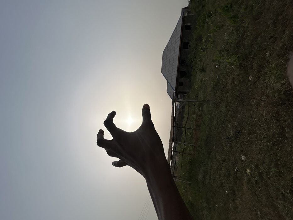 A silhouetted hand reaches toward a bright sun. The hand is dark and positioned in front of the sun, creating a dramatic contrast. In the background, a building with a sloping roof and multiple windows stands amidst grassy vegetation. The sky is a light, pale color, with a slight gradient, indicating the position of the sun. The overall scene is taken from a low angle, emphasizing the hand and its interaction with the light source.