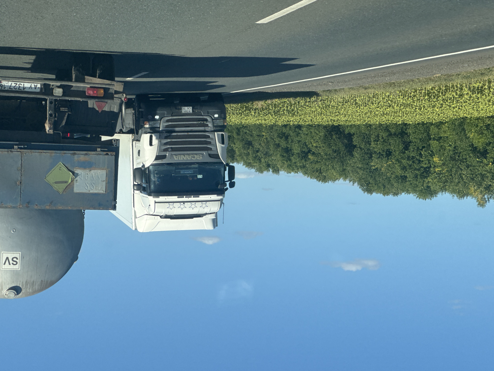 The image shows two trucks on a two-lane road. The truck in the foreground on the right is a trailer with a large cylindrical tank, likely a tanker. The truck on the left is a white Scania semi-truck. The scene appears to be rural, with a field of sunflowers on the left side of the road and a dense line of trees in the background. The sky is blue with a few scattered clouds. The road surface is asphalt with white lane markings. The license plate on the trailer appears to be AY 1327 16 RUS. The license plate on the Scania truck is H 044 CK 58. The image does not contain any people. The overall scene is a snapshot of highway travel in a rural area. There is no information within the image that indicates the location is the city of Perevolotsky, Russia.