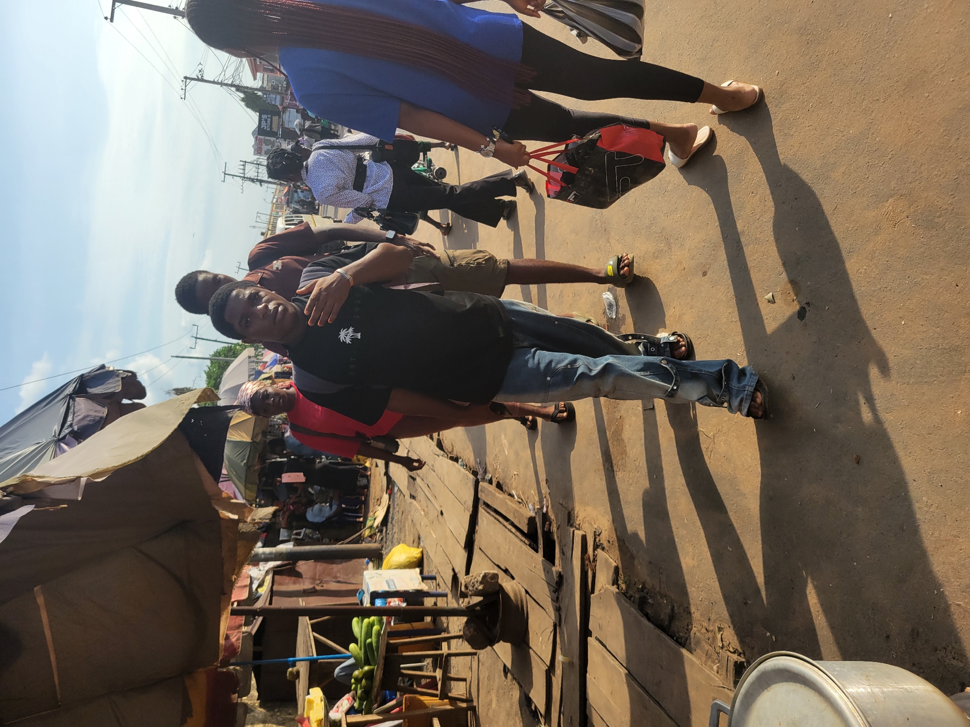 A group of people are walking on a dusty street in Egbagu, Nigeria. The scene is brightly lit by the sun, casting long shadows. In the foreground, a young man in a black t-shirt and blue jeans walks towards the camera, his left hand raised in a gesture. To his right, a woman with long braided hair, wearing a blue t-shirt and black leggings, walks away from the camera, carrying a red and black bag. Behind the young man, another man in a brown t-shirt and shorts is visible. Further back, other individuals are seen walking and standing, some under makeshift stalls with umbrellas providing shade. The environment appears to be a market, with wooden planks forming a makeshift walkway and various goods, including cucumbers, displayed on tables. Overhead wires and poles are visible in the background, suggesting an urban setting. The ground is a mix of dirt and concrete.