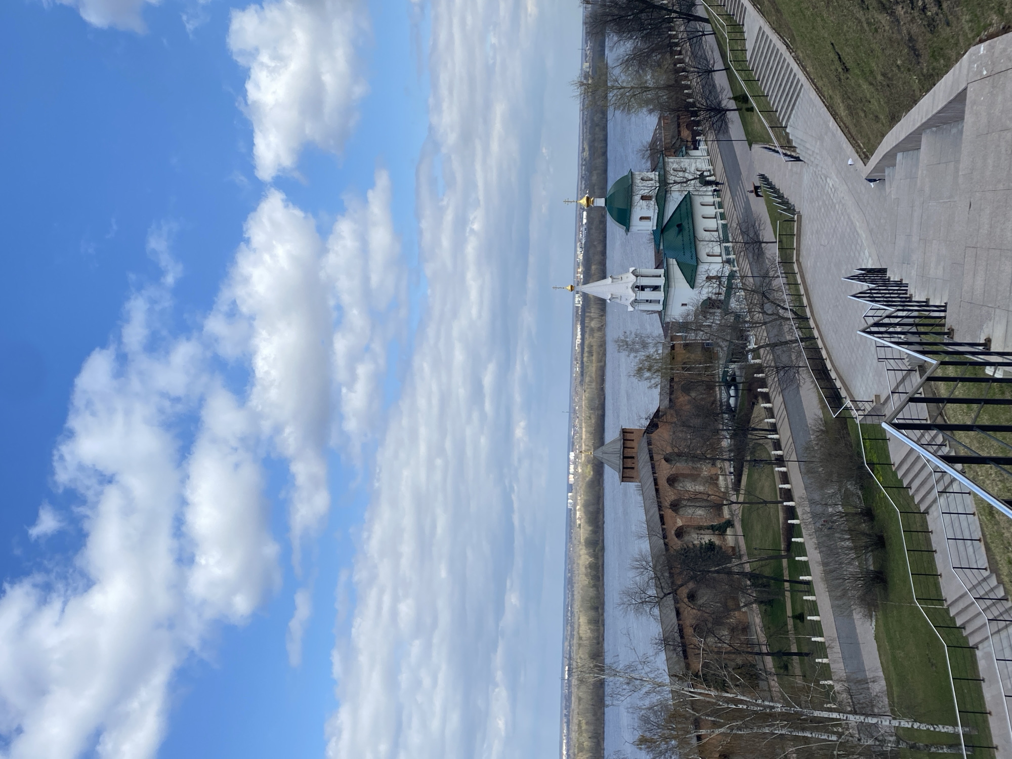This panoramic photograph, captured from an elevated viewpoint, showcases a scenic riverfront area in Nizhny Novgorod, Russia. The sky, a vibrant blue with scattered white cumulus clouds, dominates the upper portion of the image. Below, a wide river stretches across the mid-ground, its surface calm, with a distant treeline and faint structures visible on the opposite bank.
The lower half of the scene features a blend of historical architecture and modern urban landscaping. On the left, a prominent white Russian Orthodox church with multiple green onion domes and golden crosses stands gracefully near the river. To its left, partially obscured by trees, is an old red-brick fortified wall or structure, possibly part of a Kremlin, characterized by arched openings and a wooden tower.
A paved promenade, lined with bare trees, benches, and small bollards or streetlights, extends along the riverbank. A few small, indistinct figures are visible on the walkway, engaged in casual strolling. To the right, a large concrete staircase with black metal railings descends into the foreground, leading towards the promenade. Patches of green grass are interspersed along the paths and slopes.
The scene depicts a calm, clear daytime, likely in early spring due to the bare trees, with bright sunlight illuminating the historical buildings and river. There is no visible text in the image.