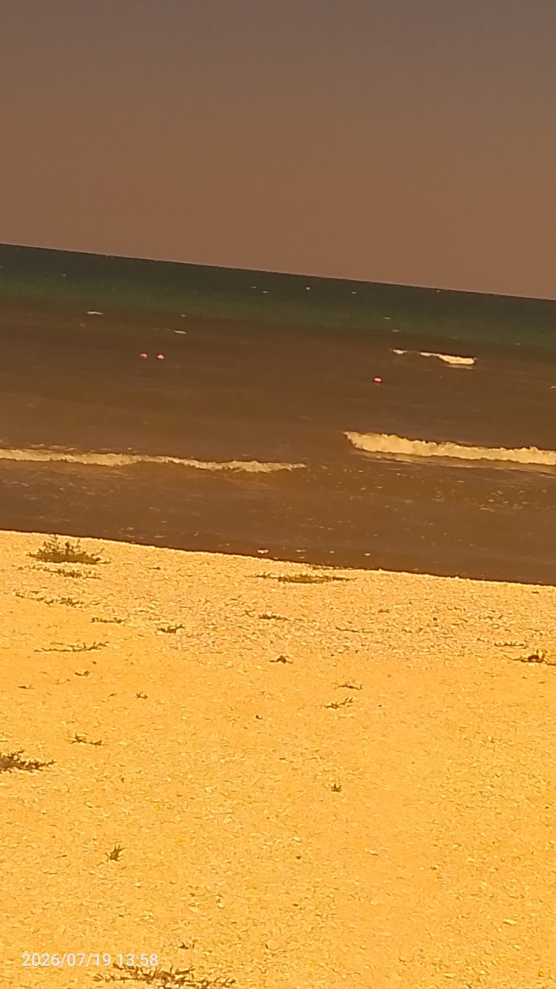 The image depicts a beach scene. The foreground is dominated by a sandy beach with sparse vegetation. The middle ground features the ocean with small waves breaking near the shore. In the distance, the sea is a darker color, and a hazy, orange sky is visible at the top of the frame. There are a few small, red objects visible in the water, possibly buoys or debris. The timestamp at the bottom left corner indicates the image was taken on 2026/07/19 at 13:58. The location is identified as Мысовское сельское поселение, Ukraine.