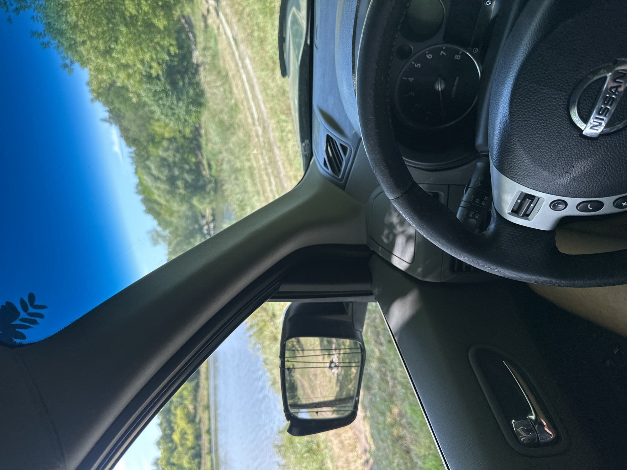 The image is taken from inside a Nissan vehicle, looking out the driver's side window and at the dashboard. The car appears to be parked on a grassy area next to a body of water. The water is calm and reflects the blue sky. Trees and greenery line the opposite bank of the water, with a dirt road visible winding through the landscape. In the side mirror, fishing rods are visible, suggesting the occupants might be fishing. The interior of the car shows the steering wheel with the Nissan logo and part of the dashboard with instruments. The scene is set in a natural outdoor environment, with the blue sky and lush vegetation suggesting a sunny day. The location is identified as Makiv, Ukraine.