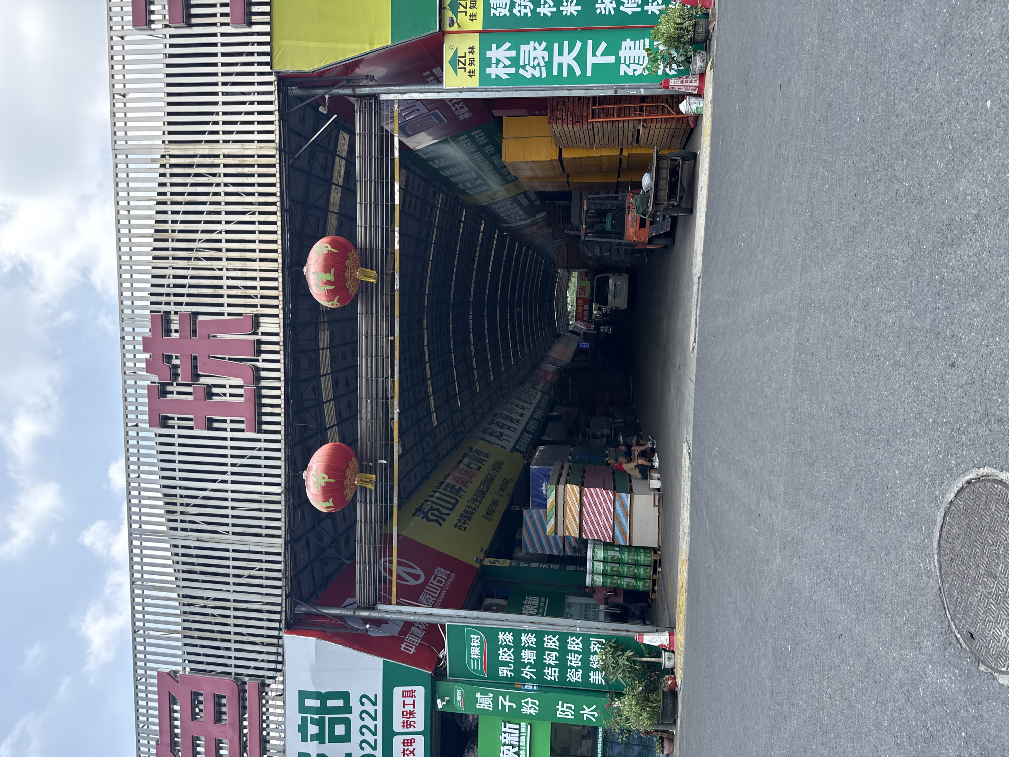 This media depicts a bustling outdoor-indoor market or warehouse specializing in building materials, likely located in Tianhe District, China, during the daytime under a clear sky with scattered clouds. The viewer is positioned on an asphalt road looking into a long, covered aisle lined with various products.
Two decorative red Chinese lanterns hang prominently from the entrance structure of the covered area. Inside, a forklift is visible, suggesting active movement of goods. Stacks of diverse building materials, including large panels, boxes, and paint cans, fill the space, reaching high towards the arched ceiling. Several individuals can be seen among the stacked merchandise, some appearing to be resting or working. The covered structure features a metal slatted facade on the left, displaying large red Chinese characters, and advertising banners hang throughout the interior. An open manhole cover is visible on the asphalt road in the foreground on the right.
Visible Chinese text includes:
- On the left facade: "部" (part of a department name) and "交电劳保工具" (Electrical Appliances, Labor Protection Tools), along with the number "2222".
- Various banners advertise brands and products such as "中国建材林泰山石" (China Building Materials Lin Taishan Stone) and "TAISHAN GYPSUM", "泰山牌局麻石膏板" (Taishan Brand Juma Gypsum Board), "林绿天下建" (Green Forest World Building), "JZL" and "佳知林" (Jiazhilin).
- Specific products like "乳胶漆" (Emulsion Paint), "外墙漆" (Exterior Wall Paint), "结构胶" (Structural Adhesive), "瓷砖胶" (Tile Adhesive), "美缝剂" (Grout/Seam Filler), "腻子粉" (Putty Powder), and "防水" (Waterproof) are listed, with the brand "三棵树" (Three Trees) appearing multiple times. Other items mentioned include "高玻" (High Glass/Fiberglass), "水保温板材" (Water Insulation Boards), "矿棉天花铝天" (Mineral wool ceiling aluminum ceiling), and "家装板" (Home Decoration Boards).