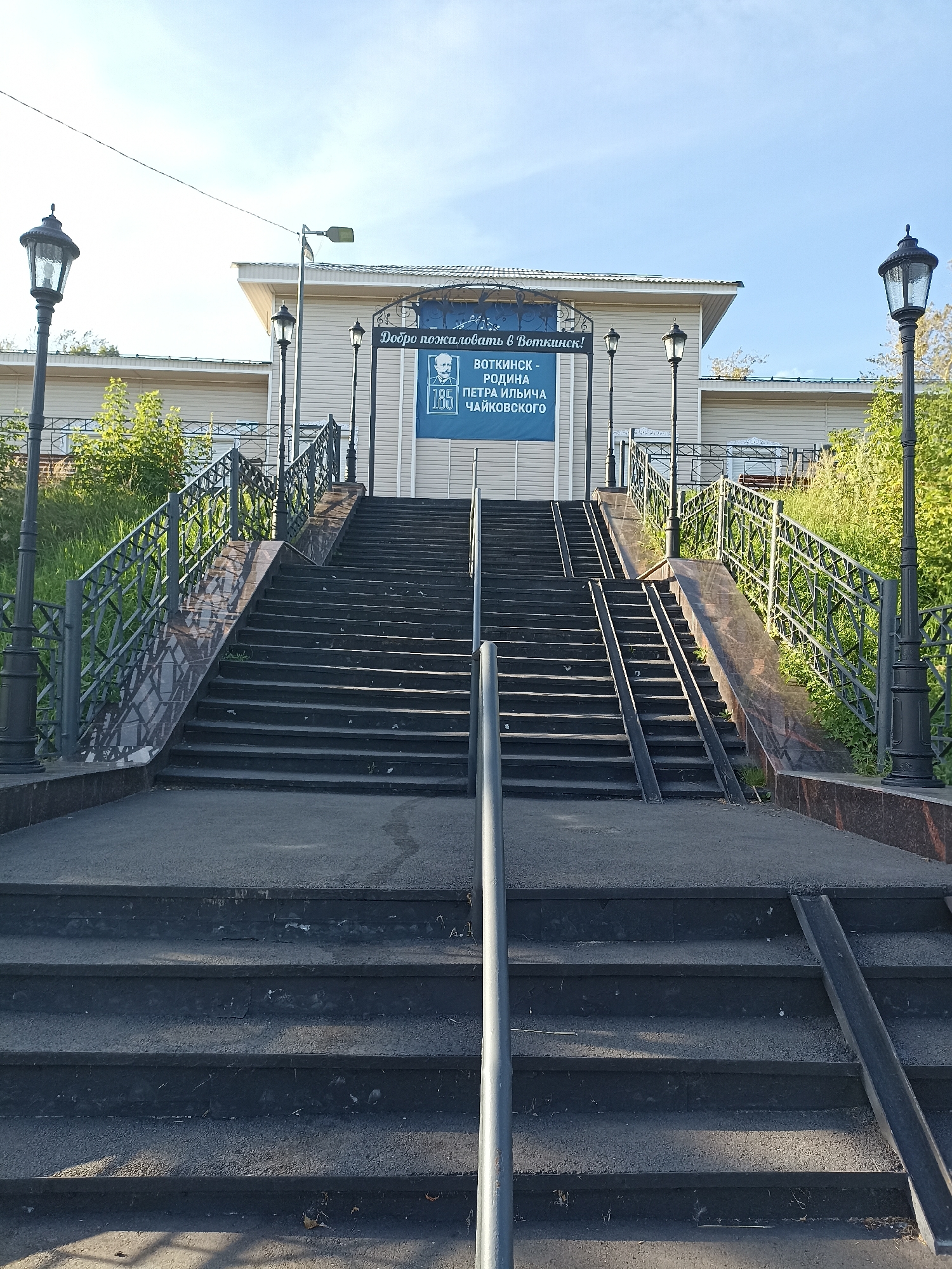 A wide stone staircase with metal railings leads up to a building. The staircase is flanked by streetlights. At the top of the staircase, a banner reads "Welcome to Votkinsk! Votkinsk - The Birthplace of Pyotr Ilyich Tchaikovsky." The building appears to be a museum or historical site. The sky is clear and blue. The scene is set in Votkinsk, Russia.