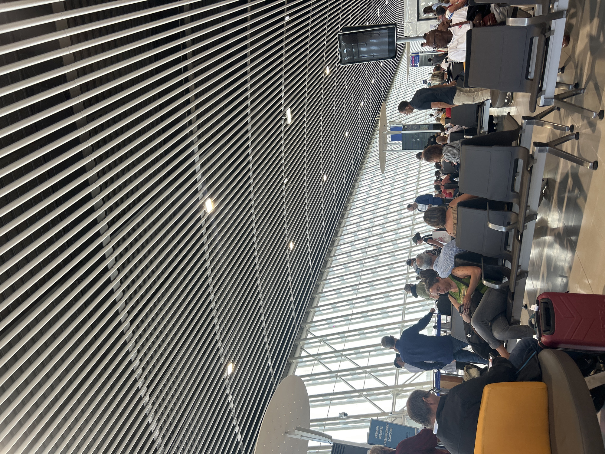 The image depicts an airport waiting area, likely in Les Abymes, France. The scene includes a number of people seated in rows of gray chairs, some with luggage nearby. A man in a blue shirt is standing, while others are seated and waiting. Overhead is a high ceiling with a distinctive pattern of parallel metal bars, possibly for ventilation or lighting, along with a partially visible glass roof. A digital information display is suspended from the ceiling. Signage is partially visible, possibly related to boarding.