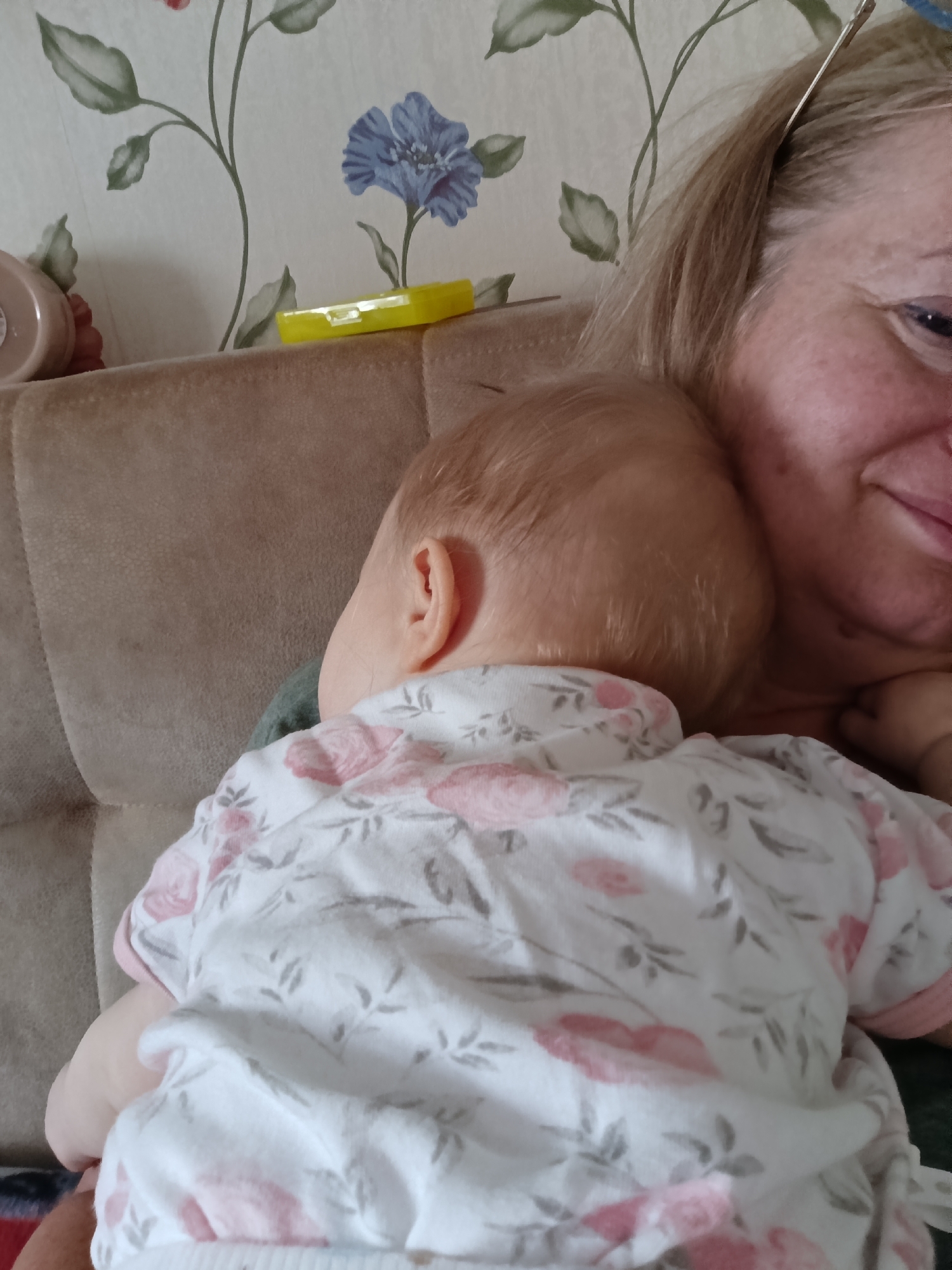 A baby in a white, floral patterned shirt is resting against the shoulder of an adult woman with blonde hair. The woman is partially visible, with a gentle smile. They are sitting on a light brown couch with a floral wallpaper background. A yellow rectangular case rests on top of the couch. The location is inferred to be Большой Букор, Russia.