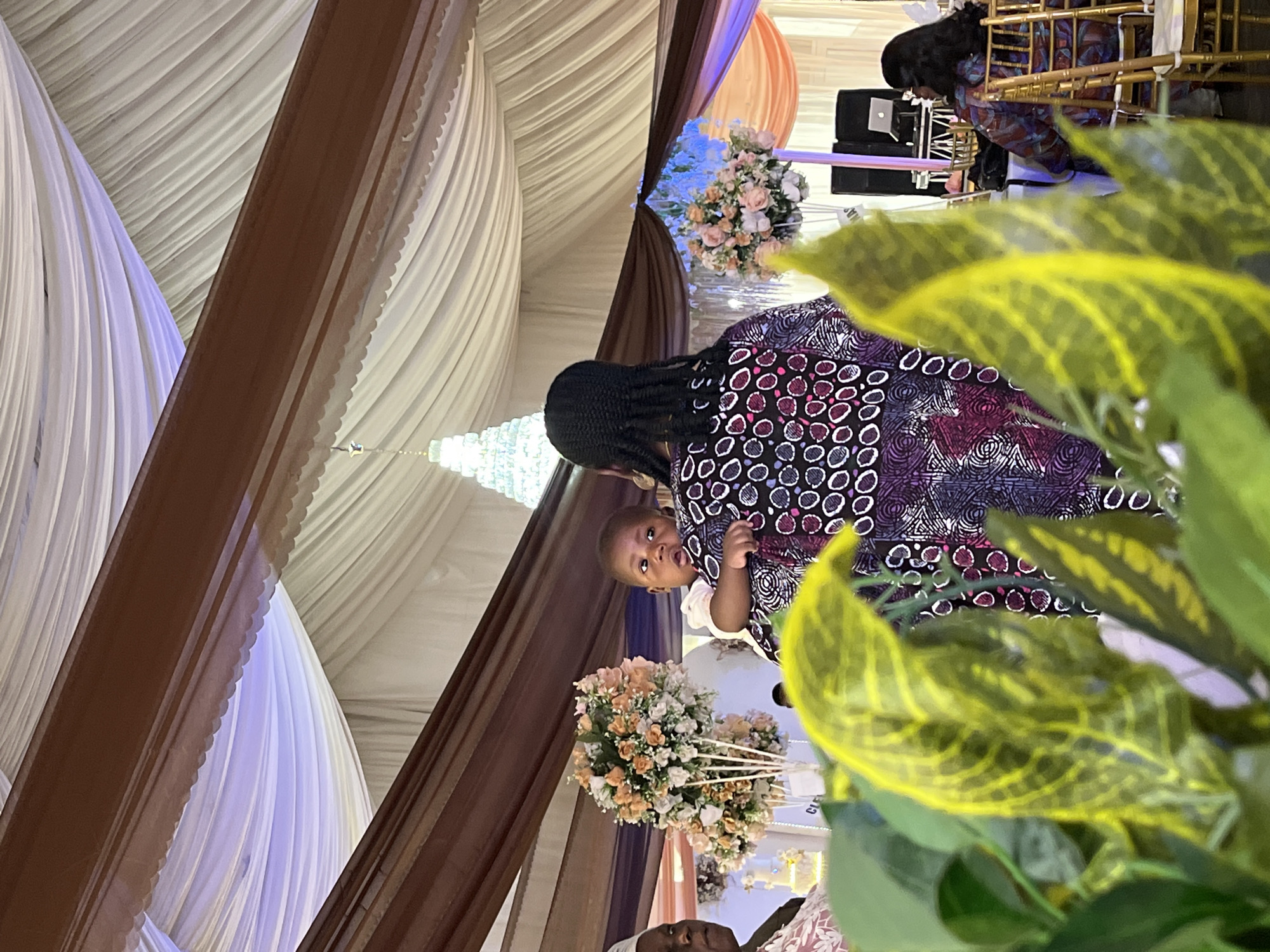 The image depicts a scene inside what appears to be a wedding or reception hall. Draped white and purple fabric hangs from the ceiling, and brown support beams are visible. A chandelier hangs overhead. A woman in a patterned dress holds a baby, and both are looking towards the camera. In the foreground is a potted plant with yellow and green leaves. In the background are flowers and what appears to be a person sitting in a chair in front of a laptop. The image was taken in Abuja, Nigeria.