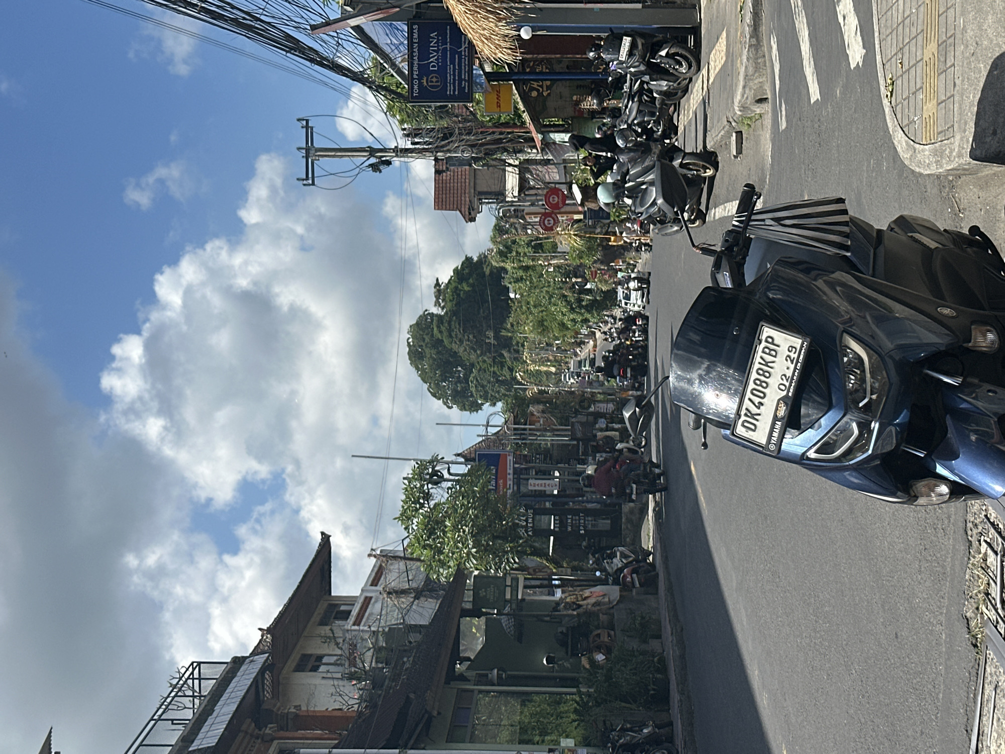 This is a medium shot, eye-level photograph taken on a street in the Ubud District, Indonesia. The scene depicts a busy street with numerous motorcycles parked along the right side and traffic moving in the distance. In the foreground, a dark blue motorcycle is parked facing the viewer, with its license plate clearly visible. The license plate reads "DK4088KBP" and has additional text that is partially obscured. The background shows a row of buildings with shops and signs, interspersed with trees. The sky is a bright blue with fluffy white clouds, and overhead, there are many electrical wires and poles. The overall impression is of a vibrant, bustling street scene in a tropical urban environment.