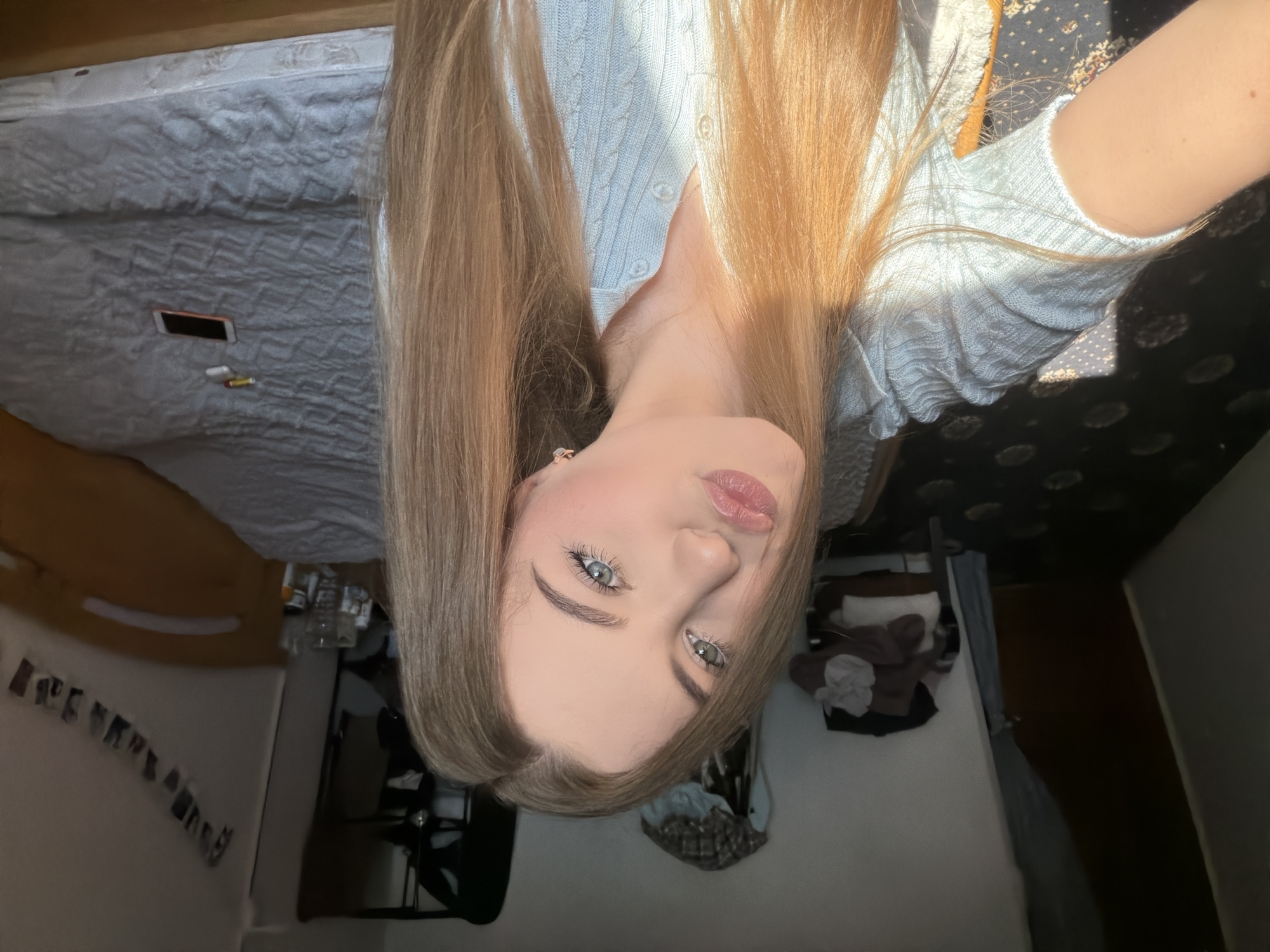 In a bedroom in Dnipro, Ukraine, a young woman with blonde hair and blue eyes is taking a selfie. She is wearing a light blue collared shirt with a cable knit pattern and has her lips pursed. Sunlight streams in from the left, illuminating her face and hair, and casting shadows on the dark carpeted floor.
In the background, a bed with a textured grey comforter is visible, with a smartphone and what appear to be a pack of cigarettes and a lighter on it. To the left, there is a closet or wardrobe with clothes hanging inside, and a chair with some folded clothes on it. A string of photos is hung on the wall above the bed. The overall impression is that of a casual, personal space.