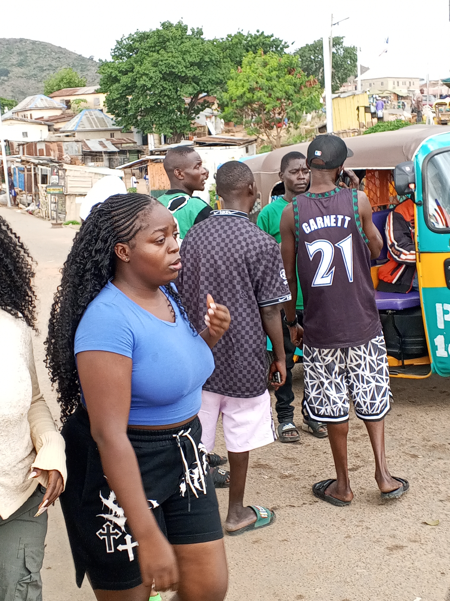 Gwong, Nigeria: A medium shot depicts a group of people on a street. In the foreground, a woman with braided hair wears a blue top and black shorts with white crosses on the left leg. Adjacent to her on the left is part of another person with curly hair, wearing a beige top and green pants. To the right of the first woman is a group of four men. The first man is wearing a checkered black and purple shirt and pink shorts, the second man is wearing a green top, the third man is partially visible, and the fourth man is wearing a 'Garnett 21' jersey and patterned black and white shorts. All four men are wearing sandals. To the right of the men is a green auto rickshaw. In the background, there are houses and trees. A mountain is visible in the far background.
