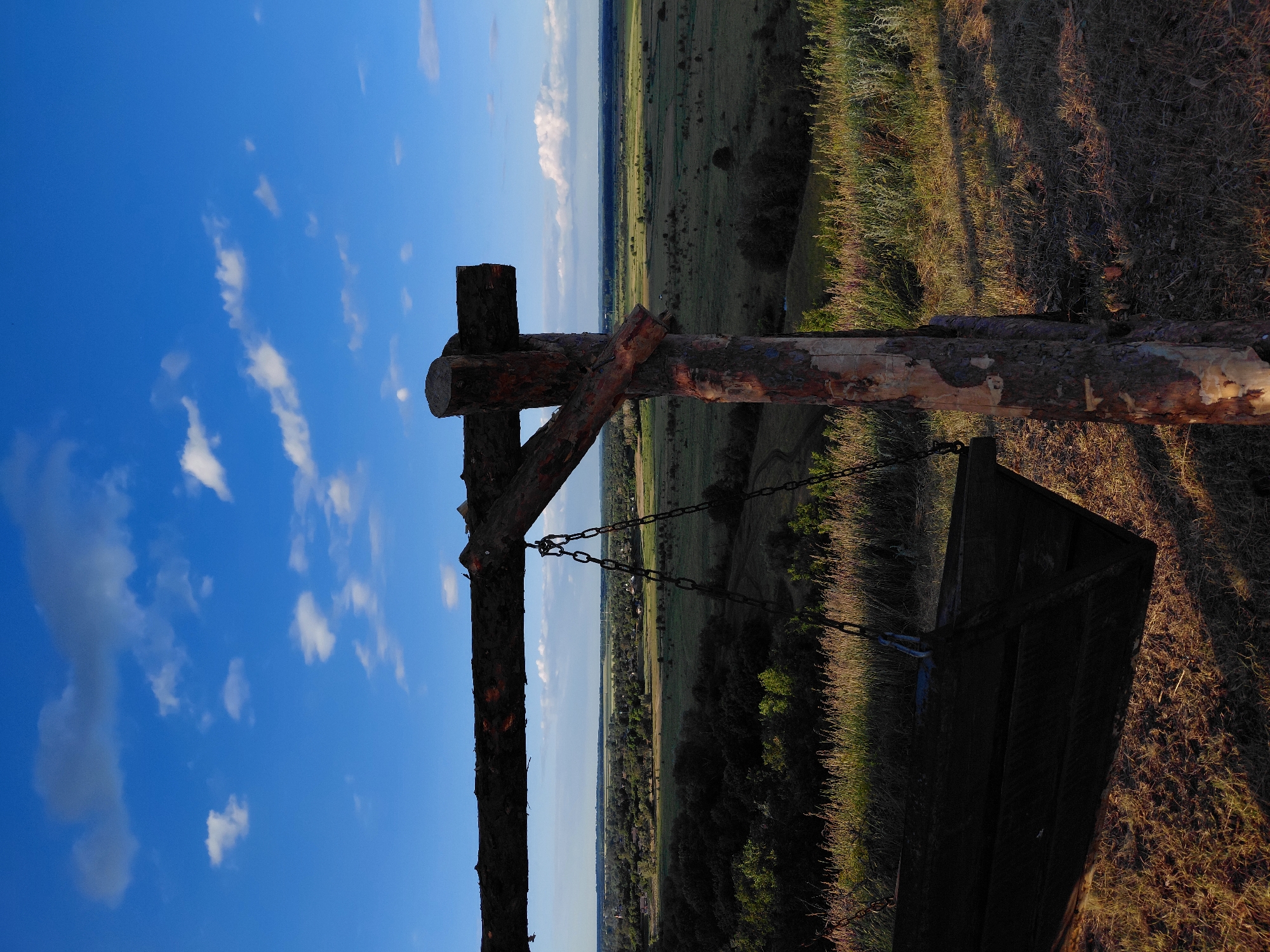 A wooden swing set is in the foreground, made of rough-hewn logs. The seat of the swing is dark wood and hangs from chains. The swing set is positioned on a grassy hill, with dry grass and patches of bare earth in the foreground. In the midground, there is a view of a valley filled with trees and fields, with a small town or village visible in the distance. The sky is blue with scattered white clouds. The lighting suggests it is late afternoon or early evening, with long shadows cast across the scene.