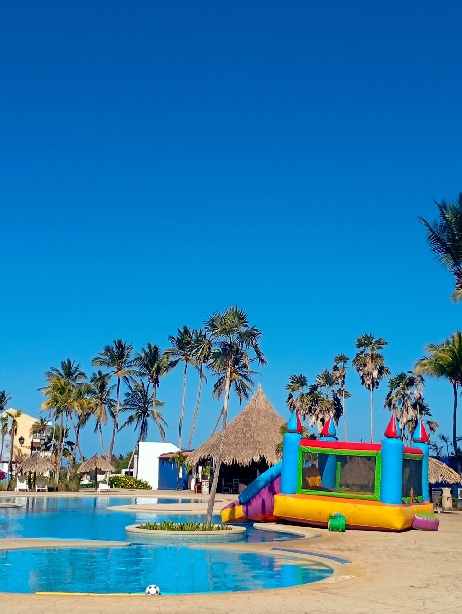 The scene depicts a sunny day at Playa El Agua, Venezuela, featuring a swimming pool complex with palm trees and thatched-roof structures in the background. A colorful inflatable bouncy castle with a slide sits beside the pool. A soccer ball is visible in the pool.