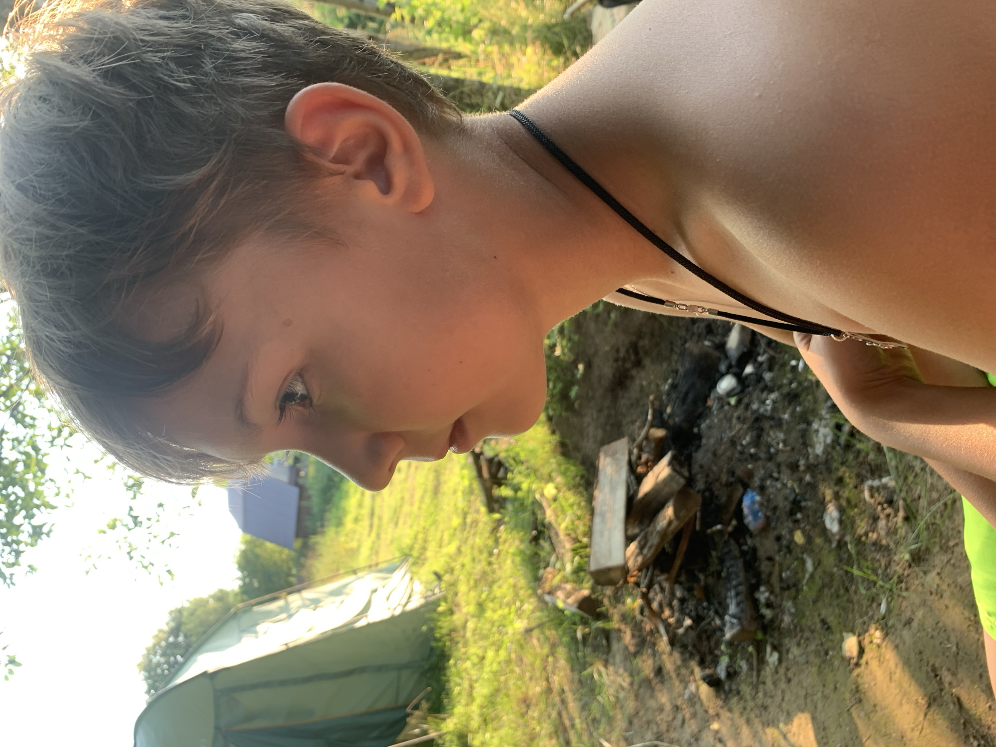 The media file is a close-up, horizontally oriented photograph of a young boy's head and shoulders, taken outdoors during daylight. The boy is looking downwards and to his left, with a neutral or slightly curious expression. Sunlight is strongly illuminating the right side of his face and neck, creating highlights and casting some shadows. He has short, light brown hair and his right ear is visible. He is wearing a thin black cord necklace.
The background is blurred but suggests a natural, rural setting. To the left and in the distance, a green tent and a dark blue structure are visible amongst trees. The ground in the foreground and mid-ground appears to be dirt with patches of grass and what looks like a small, makeshift campfire with scattered wood. The overall lighting suggests it is a sunny day, likely late afternoon or early morning due to the warm light. The presence of the tent and campfire hints at an outdoor activity, possibly camping or a backyard gathering. There is no visible text in the image. The location context provided is Zasechnyi, Russia.