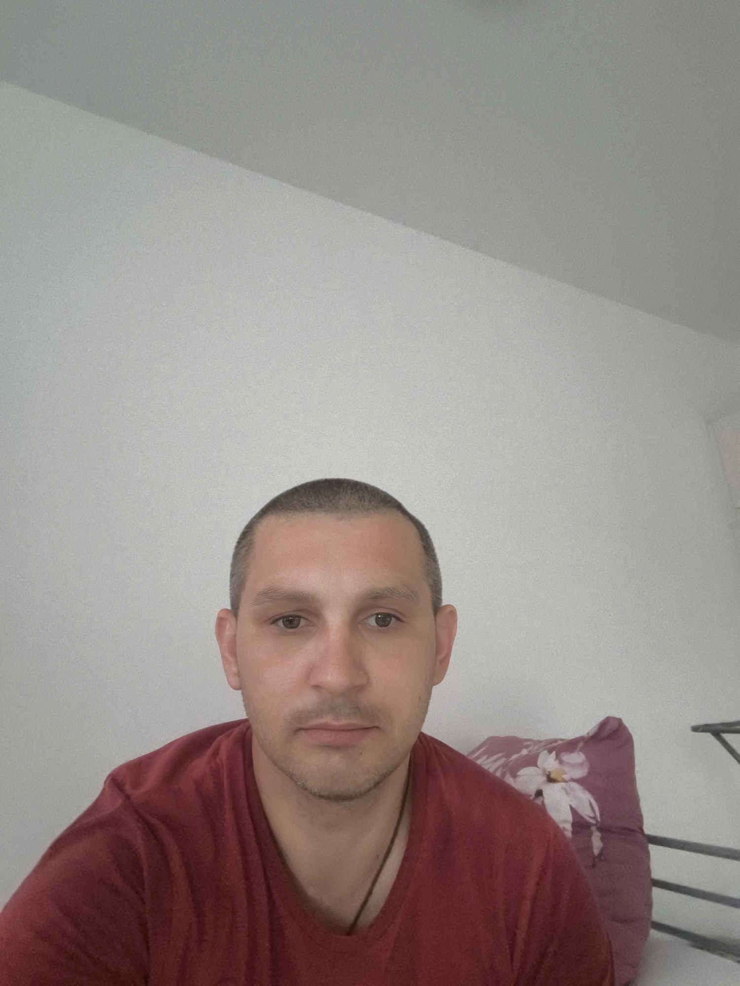 The image is a selfie of a man in his late 20s or early 30s, with short hair and a light beard, wearing a maroon t-shirt. He is looking directly at the camera with a neutral expression. Behind him, a pillow with a floral pattern is visible, and a white wall occupies the background. The scene appears to be indoors. The location in the city of Wolgast, Germany is not specified in the image or its context.