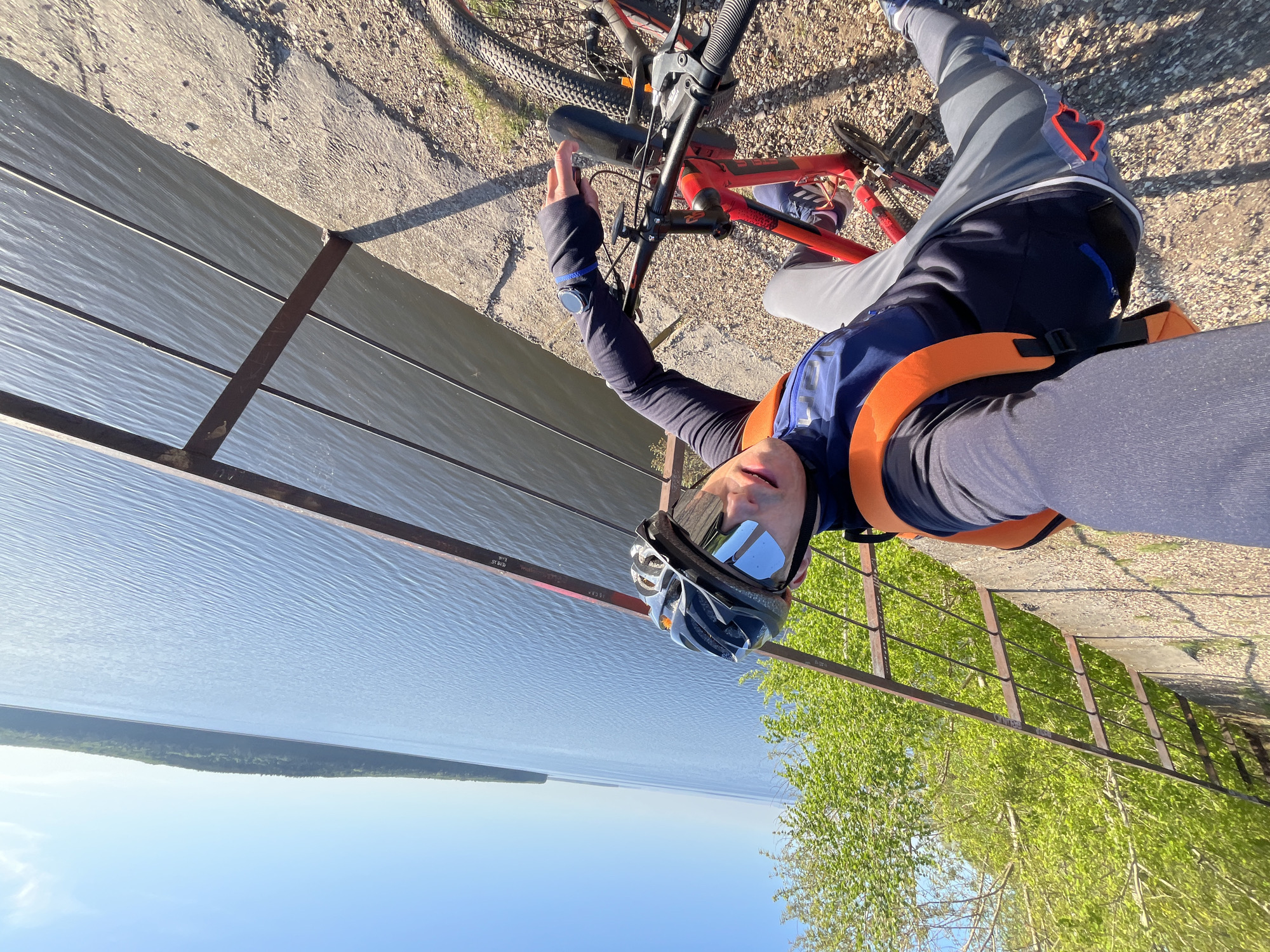 A cyclist takes a selfie on a high embankment in Chaikovsky, Russia. The cyclist, wearing a helmet and sunglasses, is on a red mountain bike. A gray metal fence runs along the embankment, overlooking a body of water with a wooded shoreline in the distance. The sky is clear blue. The cyclist's right arm is raised near the bike's handlebars. The cyclist is wearing a long-sleeved dark shirt and a visible sports watch on their left wrist.