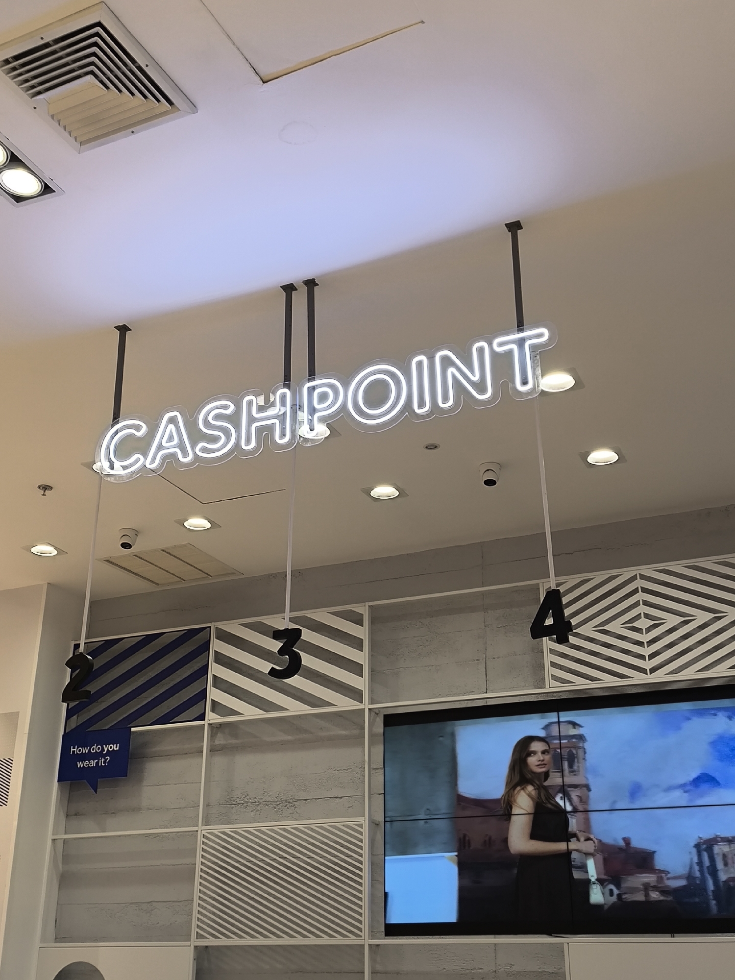 The image depicts the interior of a retail store, specifically the cash point area. The sign above reads "CASHPOINT" in neon lights. Below the sign are numbered queuing stations marked "2," "3," and "4." Station 2 includes a sign that asks, "How do you wear it?". A large screen displaying an image of a woman in front of a building is visible at station 4. The setting suggests a modern shopping environment. The location within the city of Porlamar, Venezuela, is not discernible from the image alone.