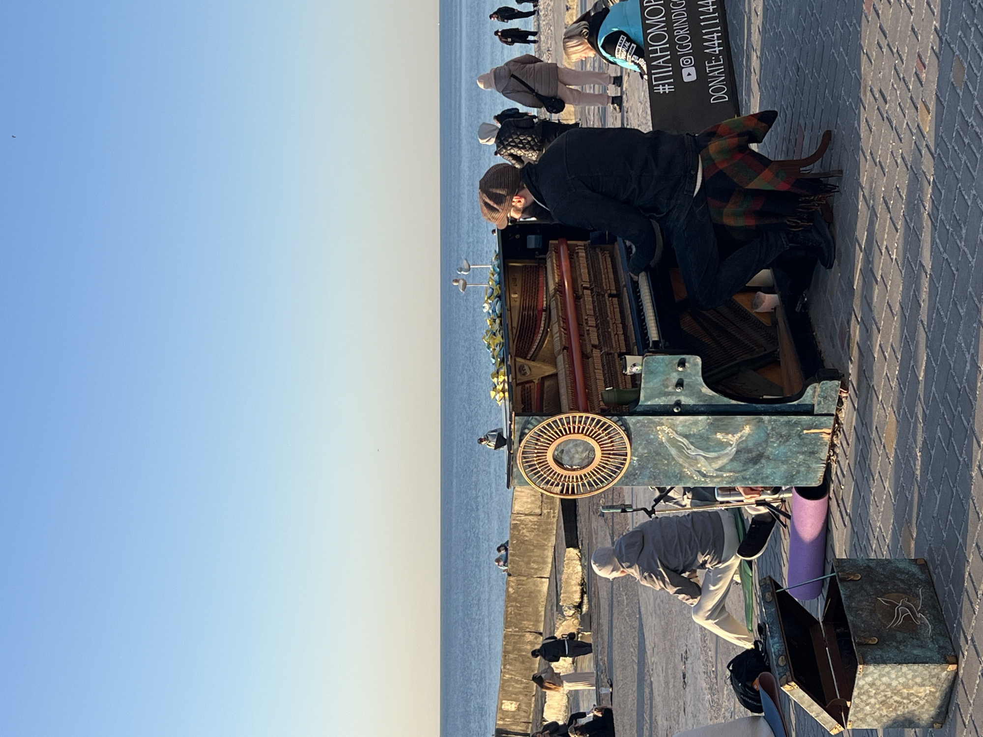 The image shows an outdoor scene on a paved area along a body of water, likely in the city of Unknown, Unknown. An old, blue piano stands in the center, its internal components exposed. A man is playing the piano, facing it, with a patterned scarf draped over one of the piano legs. Behind the piano, a sign with the hashtag "#ПІАНОМОРІ" and donation information is visible.
Another person, possibly collecting money, is crouching near the piano with an open, decorated metal box in front of them. There is a purple yoga mat leaning against the side of the piano.
Several people are standing or walking nearby, some watching the piano performance. In the background, the body of water stretches towards the horizon under a clear, light blue sky. The scene is captured in daylight.