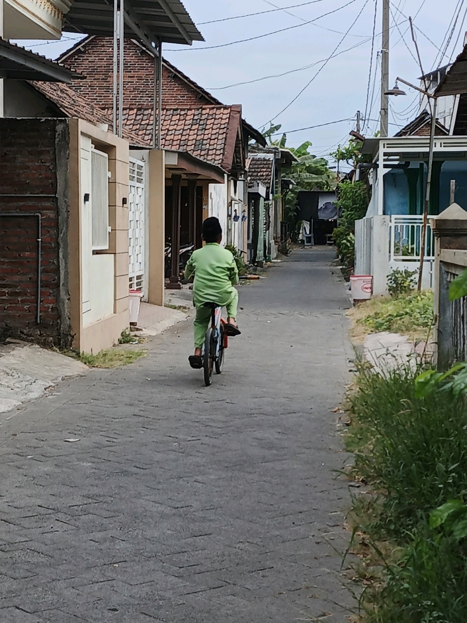In Ketegan, Indonesia, a young boy is riding a bicycle down a narrow, paved alleyway between houses. He is wearing a bright green long-sleeved shirt and pants, and a black head covering. The bicycle appears to be small, suitable for a child, and has visible spokes and pedals. The alleyway is lined with brick and plastered houses, some with tiled roofs and open doorways. There are also some trees and foliage visible. Electrical wires are strung overhead, supported by a utility pole. A red and white bucket is placed near the side of the alley. The overall scene depicts a typical residential street in the area.