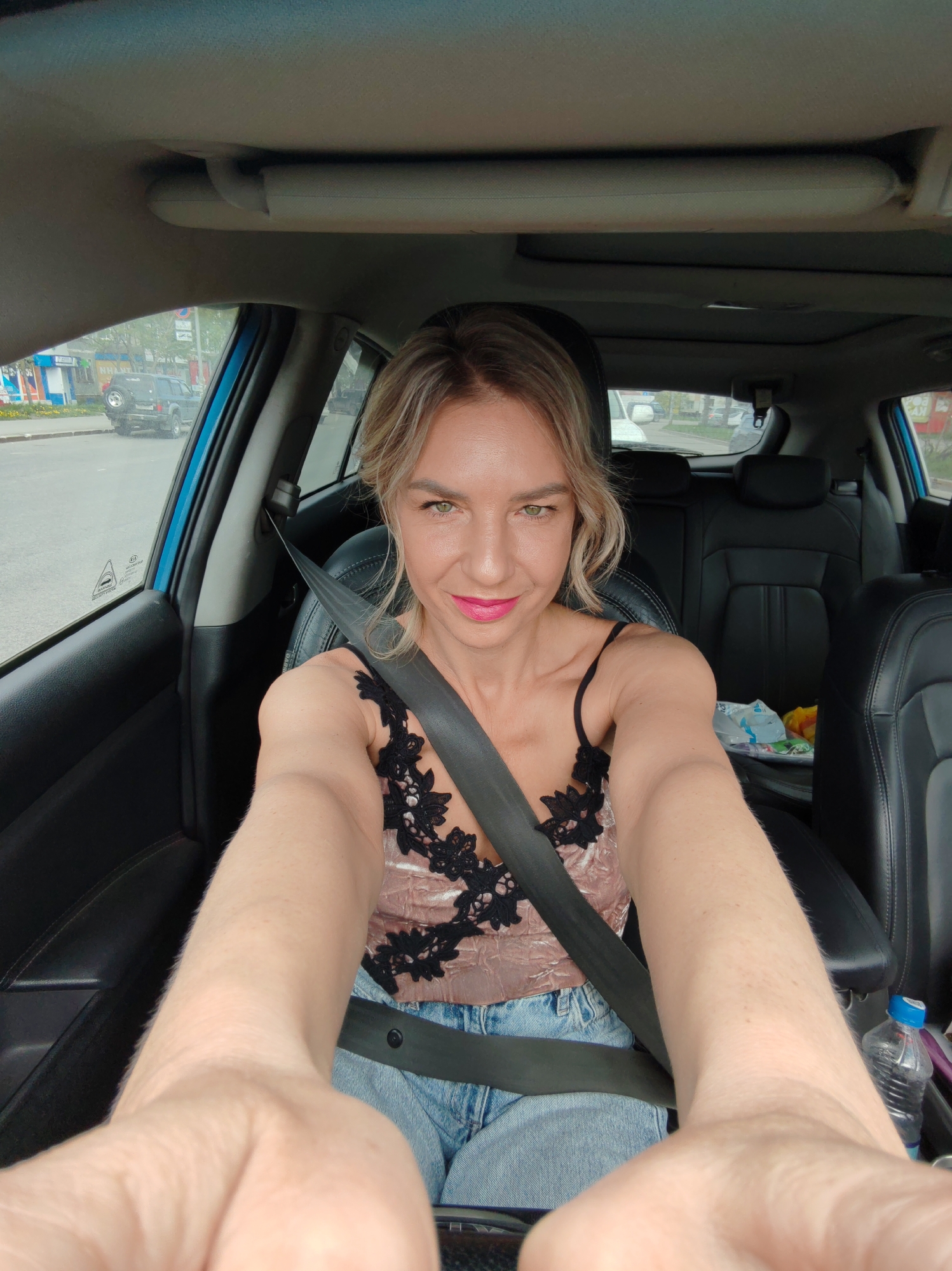 A blonde woman, wearing a pink camisole with black lace trim and blue jeans, is seated in the driver's seat of a car, taking a selfie. She is wearing a black seatbelt. The interior of the car appears to be black leather. Outside the car, the environment is a city street in Petropavlovsk-Kamchatsky, Russia, with other vehicles visible. The weather is overcast.