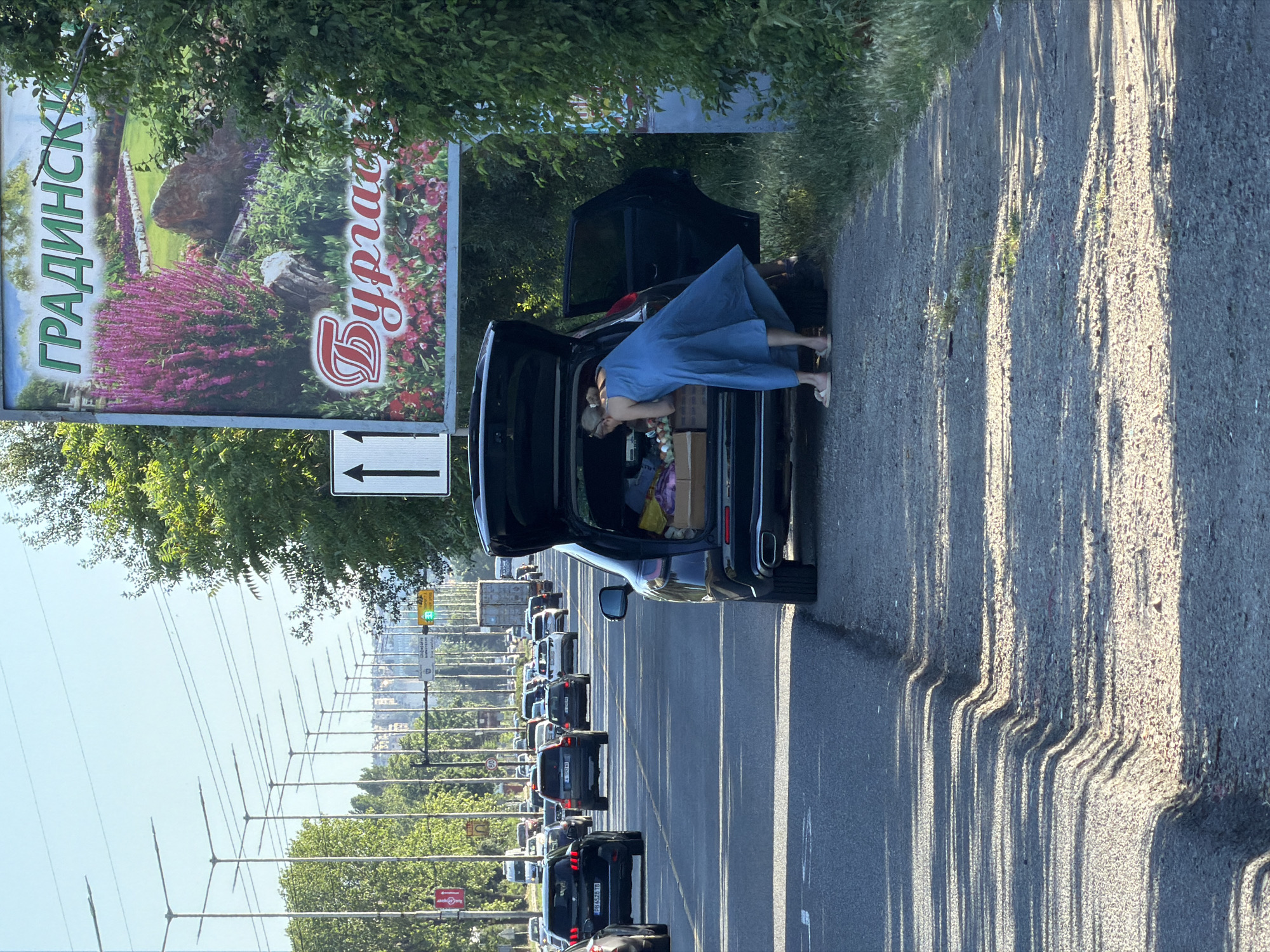 A person in a blue dress is loading groceries into the back of a dark SUV. The car is parked on the side of a busy road, with a long line of other cars stretching into the distance. The scene appears to be in Burgas, Bulgaria, as indicated by the text on a billboard that reads "БУРГАС" (Burgas) and "ГРАДИНСК" (likely part of "ГРАДИНСКИ," meaning "gardens"). The time of day is likely late afternoon, given the long shadows cast by the sun. The weather seems to be clear and sunny. The person's posture suggests they are in the process of organizing or arranging items in the trunk. The overall impression is of a routine task being performed in a public urban setting.