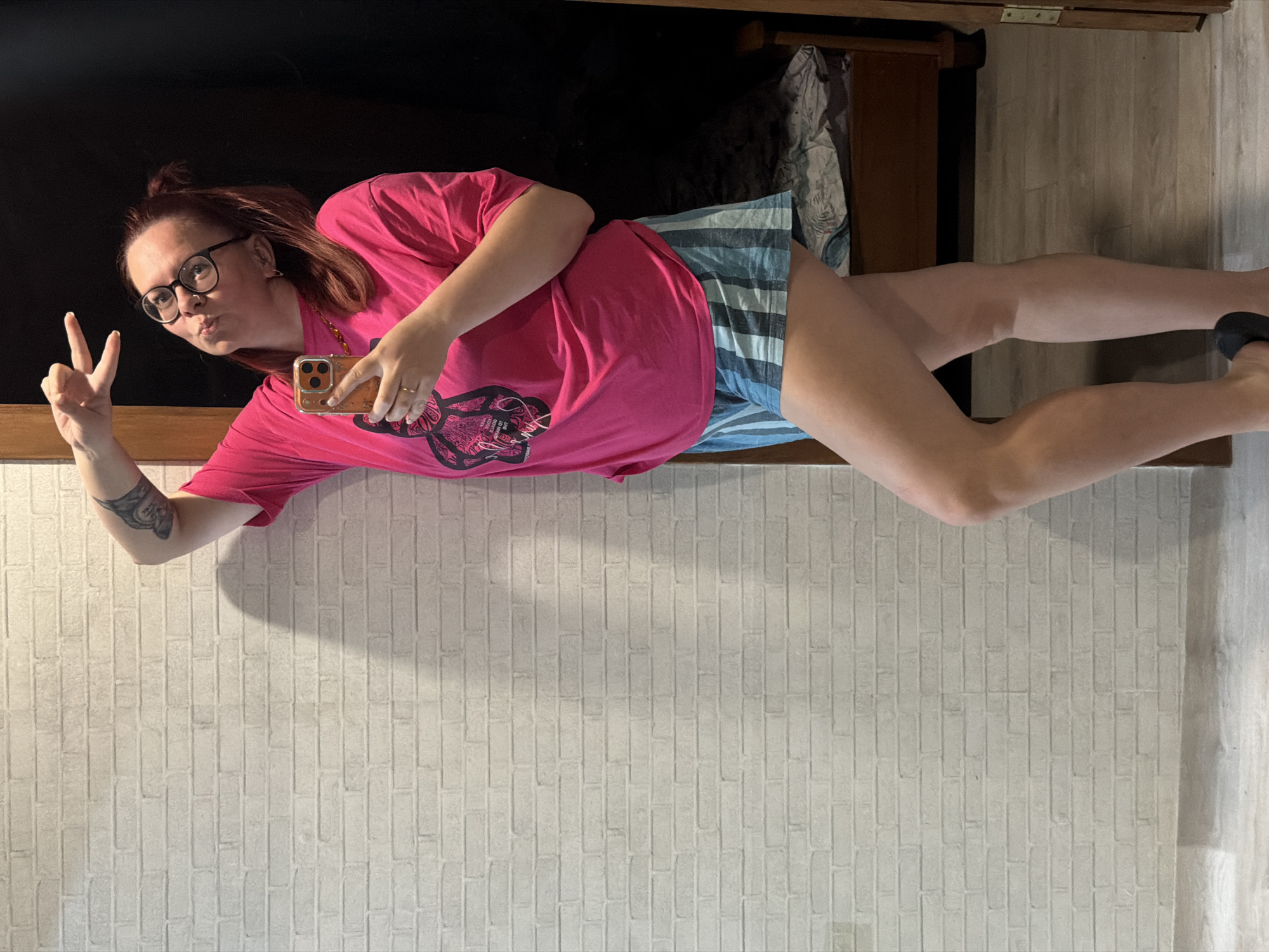 A woman stands indoors against a light-colored brick wall and a dark surface, possibly a piece of furniture. She is wearing glasses, a pink t-shirt with a graphic, and striped shorts. She is holding a smartphone in her right hand and making a peace sign with her left hand. Her hair is reddish-brown and pulled back in a bun. She has a tattoo on her left arm. The background includes a cabinet or dresser and a wooden floor. The location is Chaikovsky, Russia.