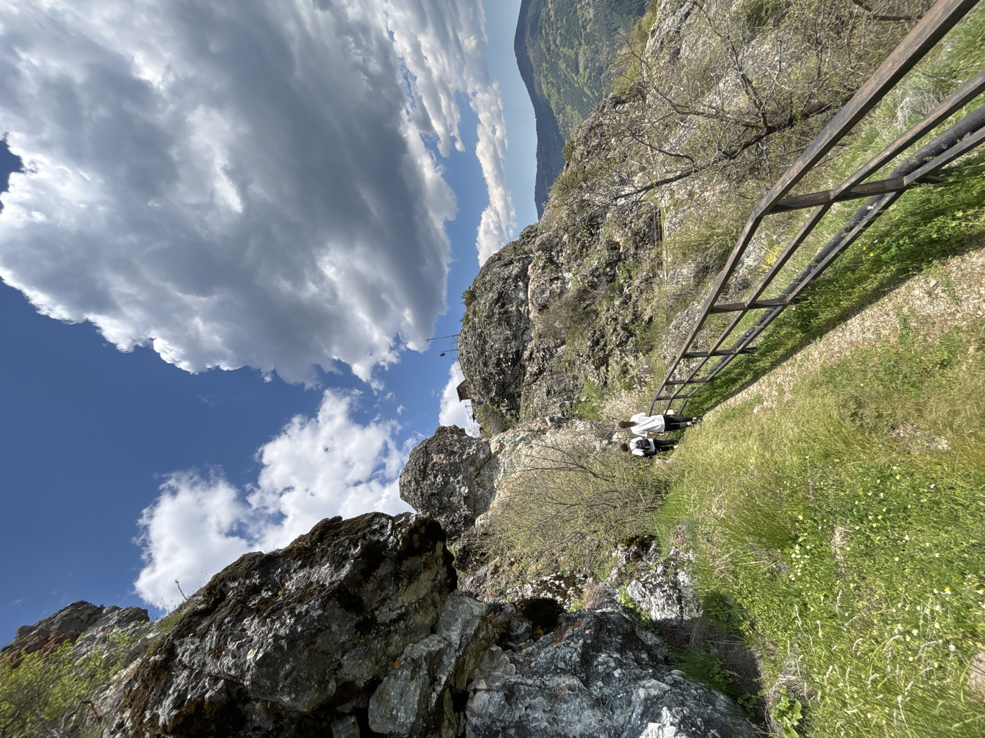 The image captures a view from a rocky, mountainous landscape near Gradat, Bulgaria, under a partly cloudy sky. The perspective is skewed, with the sky positioned towards the left. The foreground consists of craggy rocks covered with moss. A steep grassy slope descends from the center of the frame to the right. A black metal safety rail runs along the slope. Two people are visible further along the path. One person has a backpack, and both are wearing white shirts.
