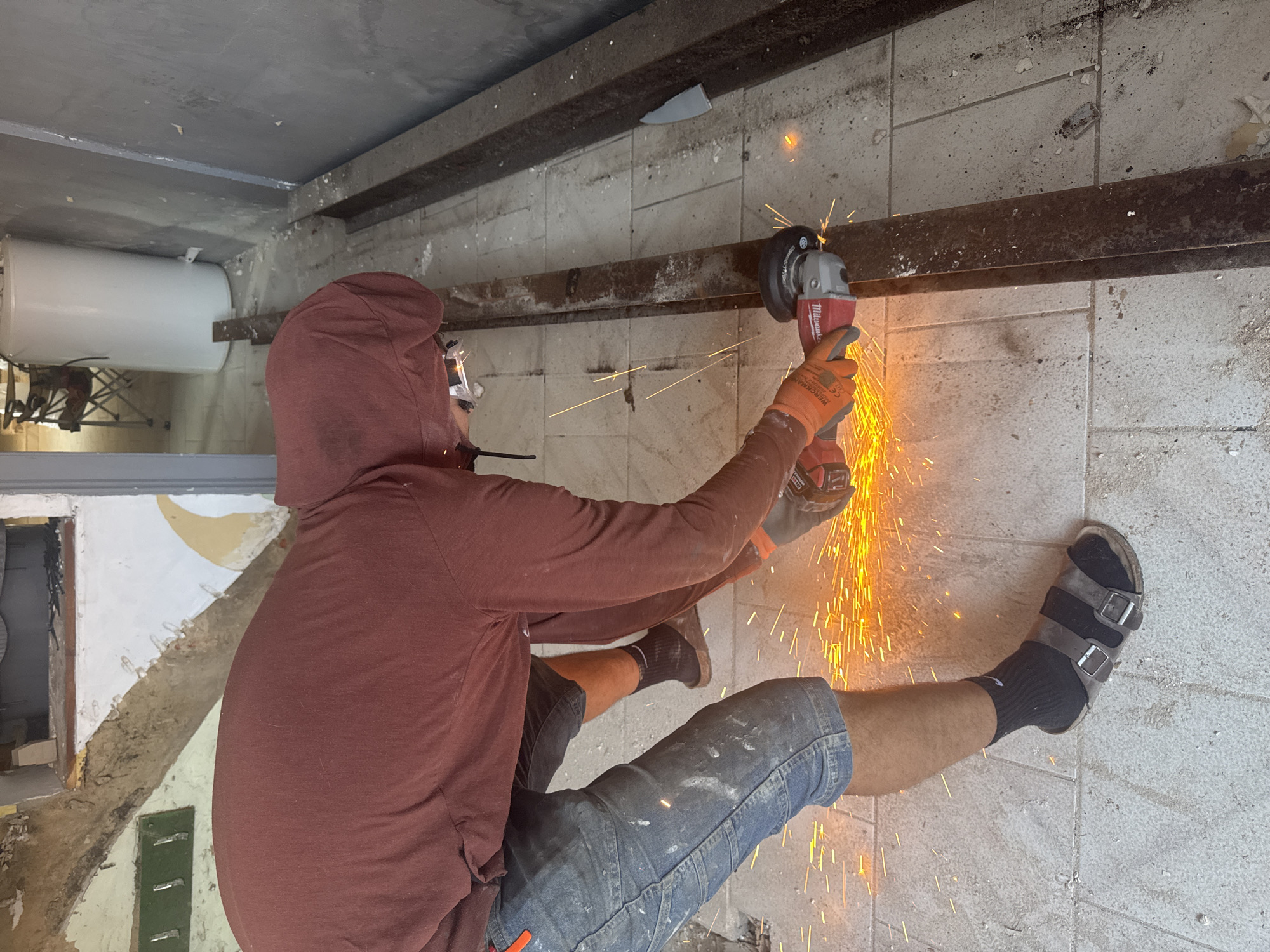The image shows a person using an angle grinder to cut a metal beam. The person is wearing a red hoodie, denim shorts, orange work gloves, and black sandals. Sparks are flying from the angle grinder. The scene appears to be an indoor construction or renovation site, with a tiled floor and exposed wall structures. There is a white water heater visible in the background, along with some furniture. The person is focused on the task of cutting the metal. The location is identified as being in Roubaix, France.
