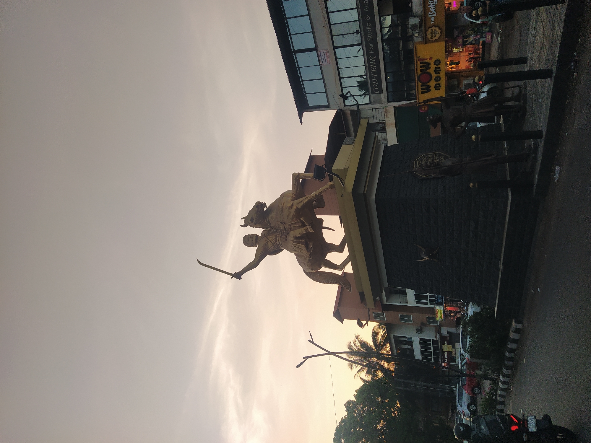 The image shows a statue of a man on horseback holding a sword, likely representing a historical figure or warrior. The statue is positioned on a pedestal. Behind the statue is a commercial area with buildings, including a hair studio and other shops with visible signage, such as "WOW! Momo." The environment suggests an urban setting, with trees and street lamps. A motorcycle is parked nearby. The sky is overcast, indicating either early morning or late evening. The location is likely somewhere in India, as the signs in the background appear to be in English with a local language. Based on visual features and signage, the city in the image is Oxel, India.