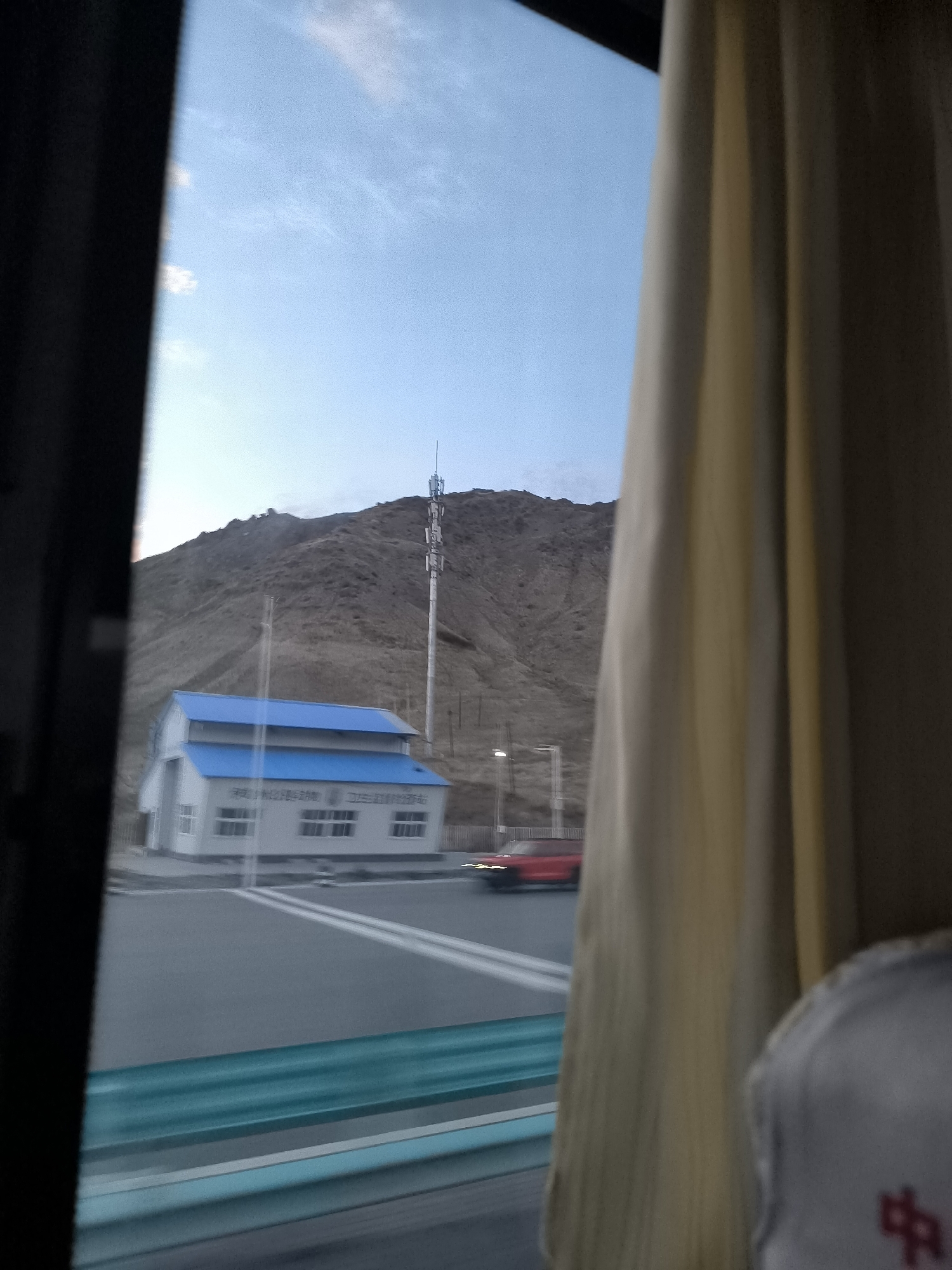 The image, captured from inside a moving vehicle, offers a view of a landscape under a clear sky with scattered clouds. In the foreground, a light-colored curtain obscures the right side of the frame, while a dark object, likely part of the vehicle's interior, is visible on the left.
The scene depicts a building with a blue roof situated on a paved road. The building appears to be white with multiple windows. A telecommunications tower stands tall behind the building on a barren, hilly terrain. A red car is seen driving on the road. The road itself is marked with white lines, and a turquoise barrier is present. The sky is a pale blue, transitioning to a lighter hue near the horizon. The overall impression is one of a somewhat remote, possibly rural or highway-side location, characteristic of areas like 果子沟牧场, China, known for its natural landscapes and infrastructure connecting different regions.