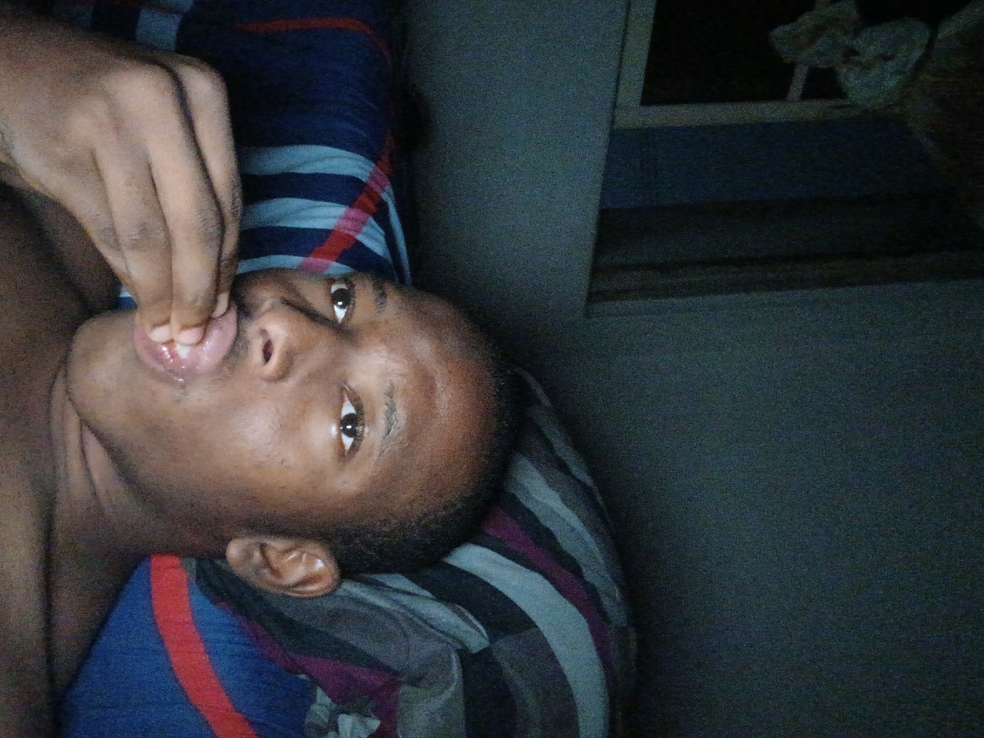 A close-up, poorly lit photograph depicts a dark-skinned, shirtless male lying on his side on a striped blanket or bedsheet. His face is turned towards the camera, and his fingers are near his mouth. The setting appears to be an indoor space with a partially visible window or frame in the background. There is no explicit indication of this being Lagos, Nigeria.