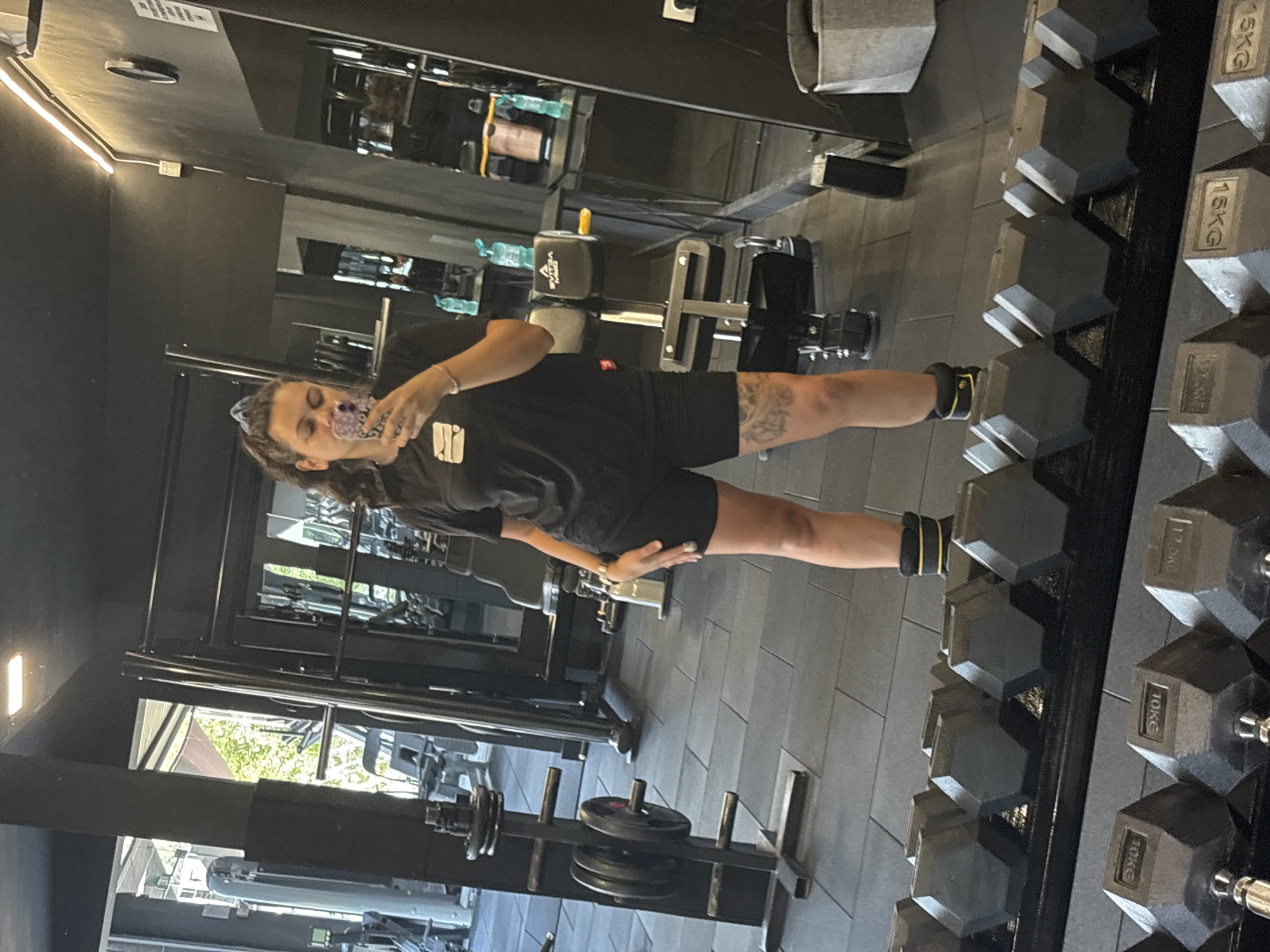 A young woman is taking a mirror selfie in a gym located in Troyan, Bulgaria. She is wearing a black t-shirt and black shorts, with black ankle weights on each leg. Her hair is dark and tied back. In the background, gym equipment such as weight racks, dumbbells, and weight machines are visible. The gym floor is made of black rubber tiles.