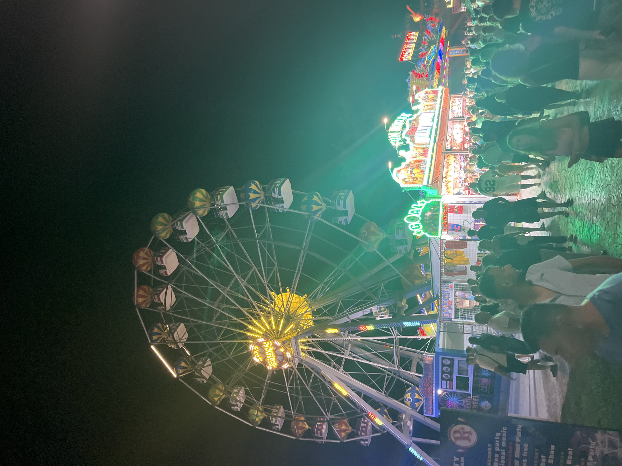 The image depicts a nighttime scene at a carnival or amusement park. A large Ferris wheel dominates the left side of the frame, illuminated with yellow and white lights. Several gondolas are visible on the wheel, some with hot air balloon designs. In the background, to the right of the Ferris wheel, is a row of illuminated carnival attractions, including a "Fun House" and other rides with flashing lights and signage. A large crowd of people, dressed in casual summer attire, are gathered and milling about in front of the attractions. The ground appears to be paved. The overall atmosphere suggests a lively evening event. Based on the context of Varna, Bulgaria, this scene is likely from the Varna Summer International Festival, which often features such attractions.