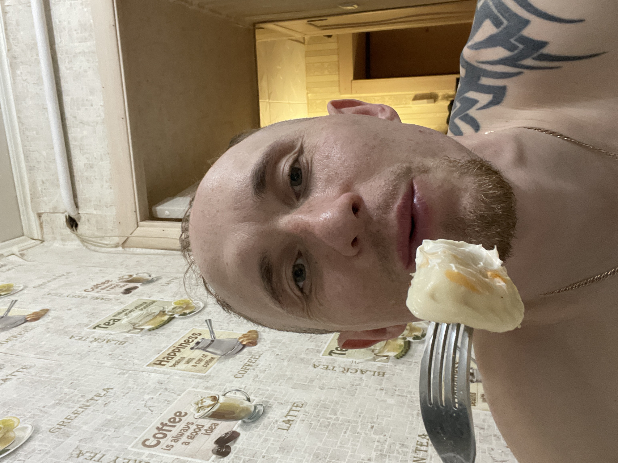 A close-up selfie shows a shirtless man with a fair complexion, a short beard, and a tattoo on his left shoulder. He is holding a fork with a large piece of food on it near his mouth. He appears to be inside a building. The background shows what seems to be a wall and shelving. The table in front of him is covered with a patterned tablecloth with designs of coffee and tea-related images and text. The text on the tablecloth includes "Coffee," "Tea," "Latte," "Black Tea," "Green Tea," "Coffee is always a good idea," and "Happiness begins with a cup of coffee." There is no visible evidence to suggest the photo was taken in Sarapul, Russia.
