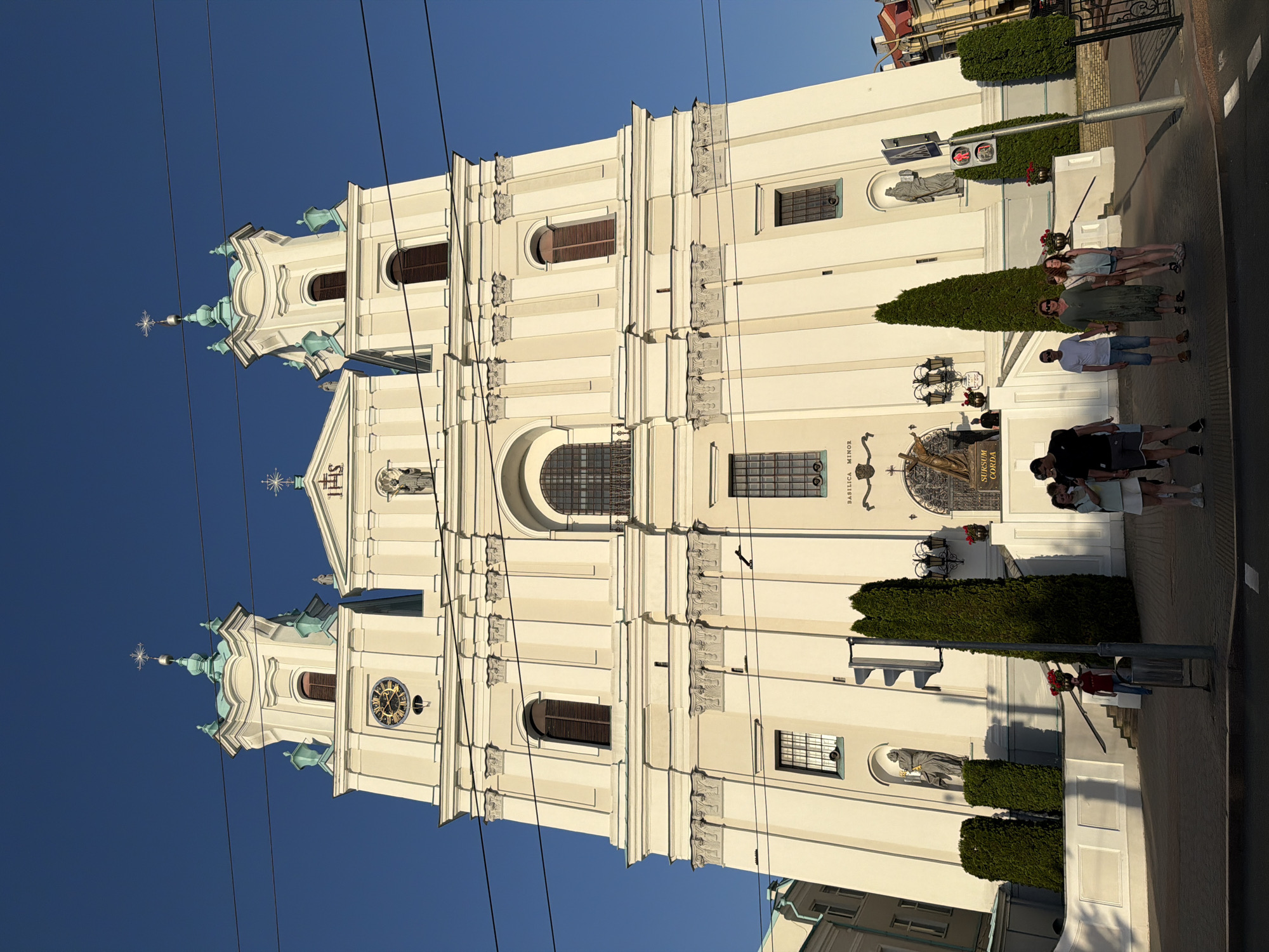 An eye-level, medium shot captures the exterior of a large, ornate white church with a bright blue sky in the background. The church features two prominent towers, each topped with a light green dome and a cross. A clock face is visible on the left tower, and the inscription "JHS" is centered above the main entrance. The facade is decorated with columns and architectural details.
In front of the church, a group of seven people are standing on a paved area. From left to right, a man wearing a black t-shirt and shorts stands with his back to the camera. Next to him, a woman in a green dress and sunglasses is facing the camera. To her right, a young woman in a blue button-down shirt and shorts is looking towards the church. A man in a white t-shirt and blue shorts stands next to the woman in green. A man in a black t-shirt and shorts is partially visible behind the young woman. Two other people are further to the right, also appearing to be looking at the church.
Several tall, slender evergreen trees are planted in front of the church. A traffic light is positioned to the left of the entrance, and a trash receptacle is near the traffic light. Red flowers are in vases on pedestals on either side of the entrance. The scene is illuminated by sunlight, casting shadows on the ground and the church. The overall impression is of a sunny day with a group of people observing a grand religious building.