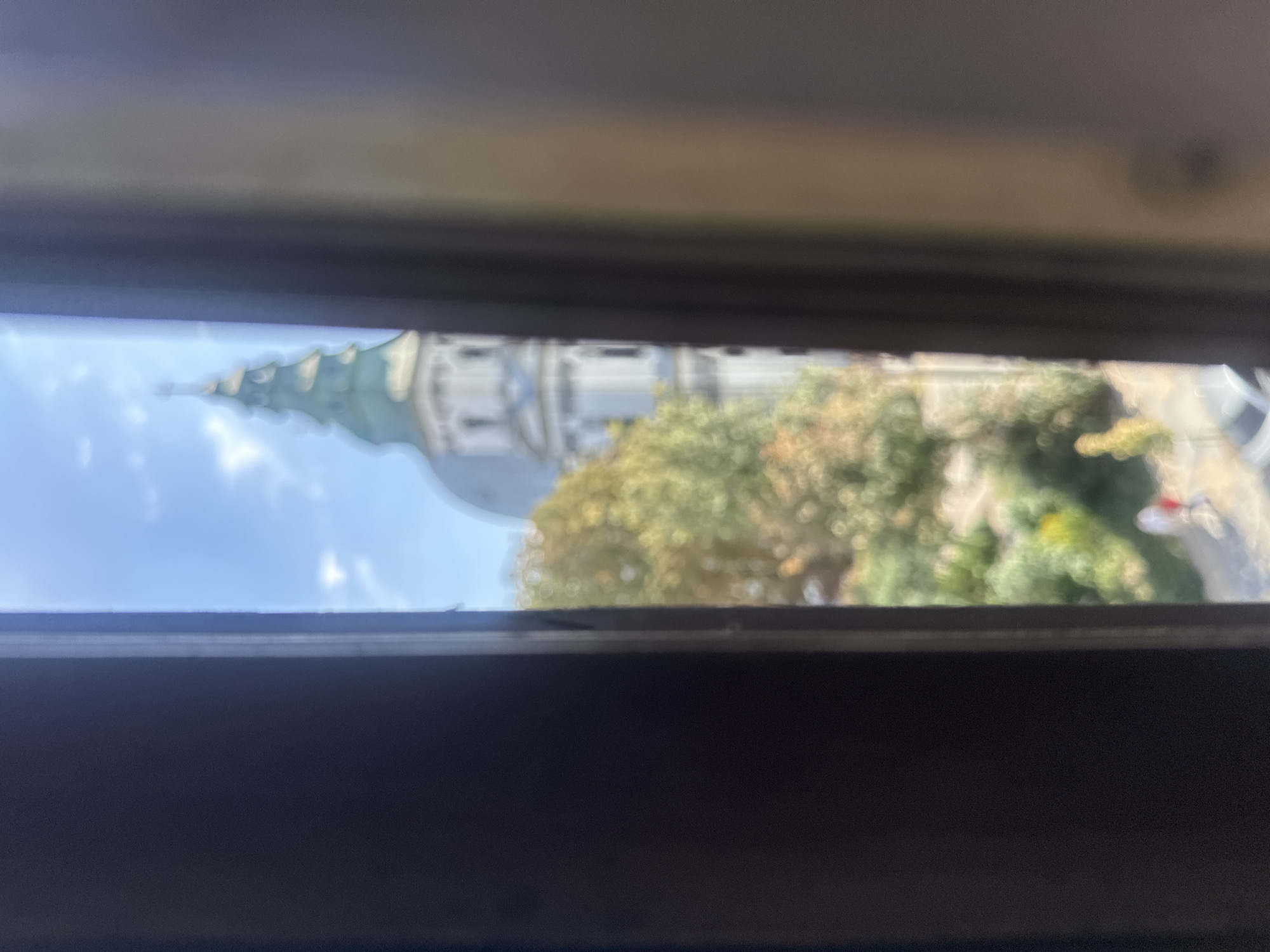 The image captures a view through a window frame in Ternopil, Ukraine. In the background, partially obscured and out of focus, is a white building with a distinctive green, multi-tiered domed roof topped with a cross. Trees with green and yellow foliage are visible in front of the building. The sky is blue with scattered white clouds. In the foreground, the dark, vertical frames of the window are prominent. A blurred pathway is visible below the trees, with a few indistinct figures walking on it.
