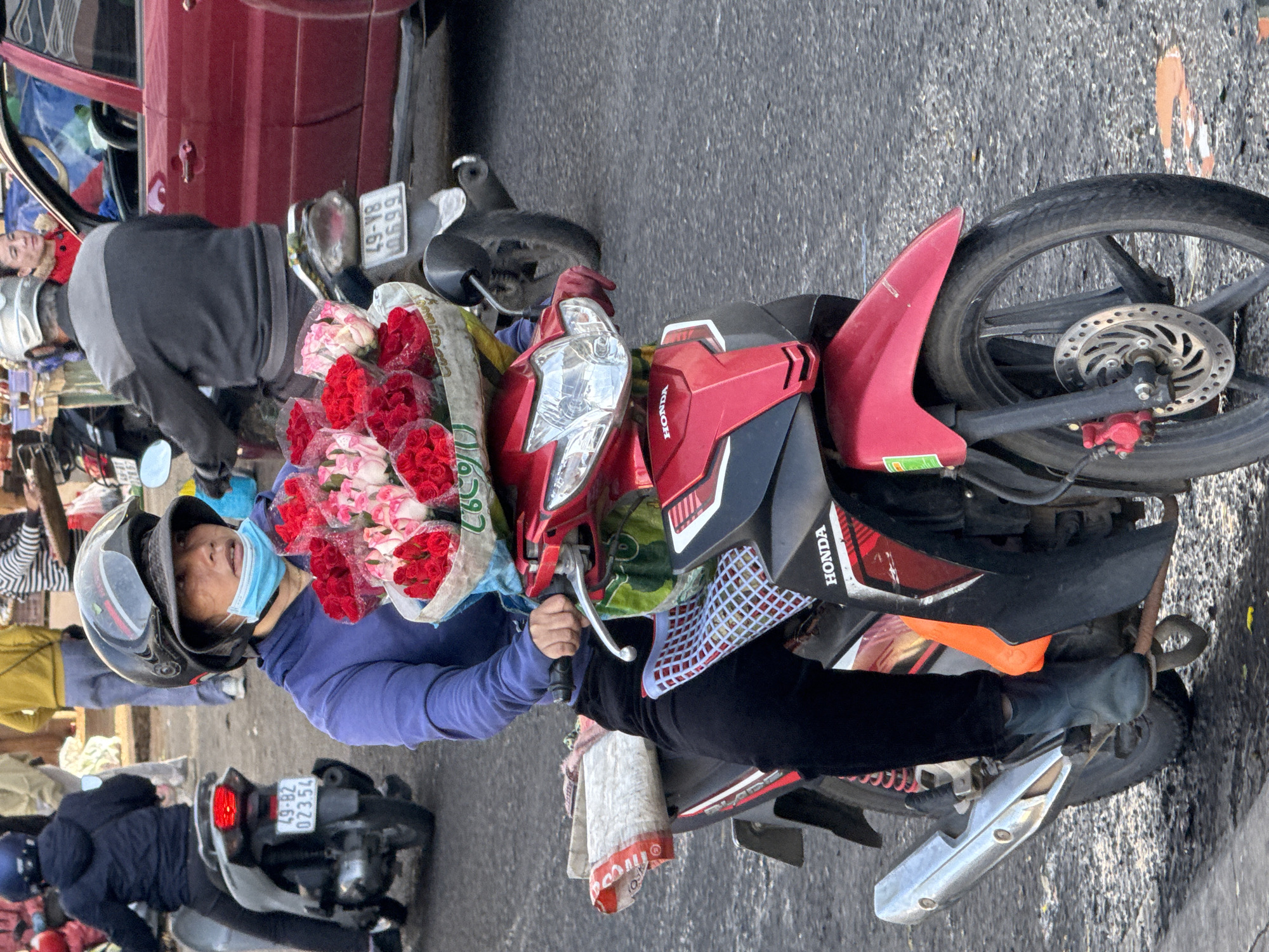 A Vietnamese woman with a blue surgical mask and a black helmet is riding a red and black Honda motorcycle in Da Lat, Vietnam. She is carrying a large bouquet of red and pink roses in front of her. The bouquet is wrapped in white and blue paper. She is wearing a purple hoodie and black pants. The motorcycle has a license plate with the number "49-B1 073.54". The background shows a blurred street scene with other vehicles and pedestrians. The road is asphalt and appears to be wet in places, indicating recent rain or a spill.