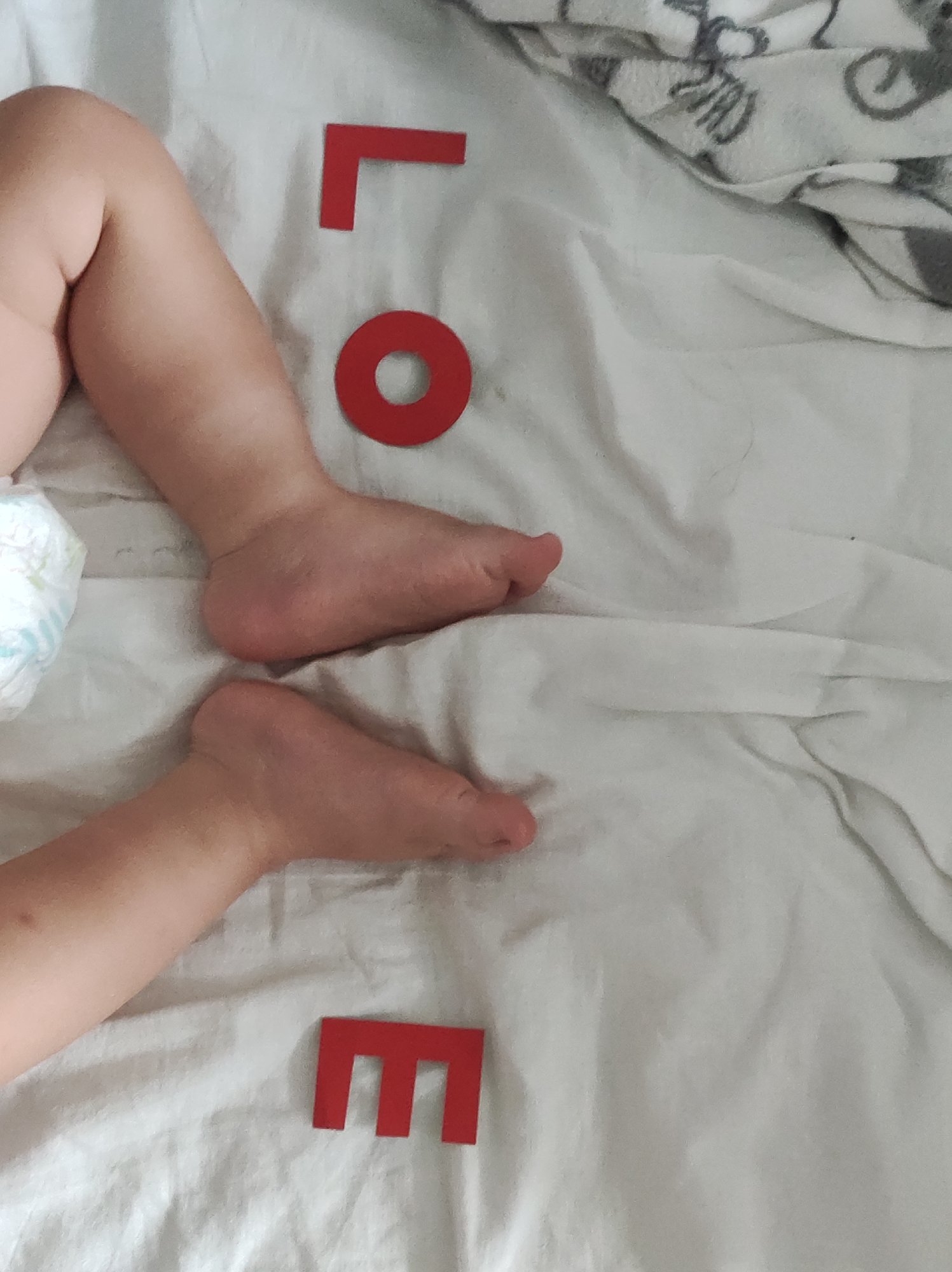 The image shows a baby's legs and feet on a white sheet. Red letters, spelling out the word "LOVE," are arranged near the baby's body. The setting appears to be a bed. There is a blanket with grey animal print design in the upper right corner. The location, Chaikovsky, Russia, cannot be confirmed from the image alone.