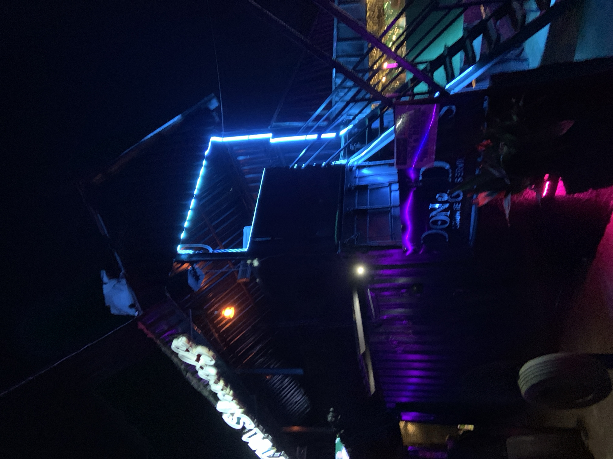 This image depicts a nighttime scene at a venue, possibly a bar or restaurant, in Gura Top, Nigeria. The structure appears to be multi-story, with a prominent set of outdoor stairs illuminated by bright blue neon lights. Purple neon lights are visible along the lower level. A sign with white lettering, partially obscured, is mounted on the upper part of the building, and another sign with "DONG" and what appears to be "SERVICE" is visible on a dark surface on the lower level, also lit by neon. The overall aesthetic is one of a vibrant nightlife establishment. The environment is dark, characteristic of nighttime, with the artificial lighting creating a colorful and somewhat dramatic atmosphere. No people are clearly visible in the frame.