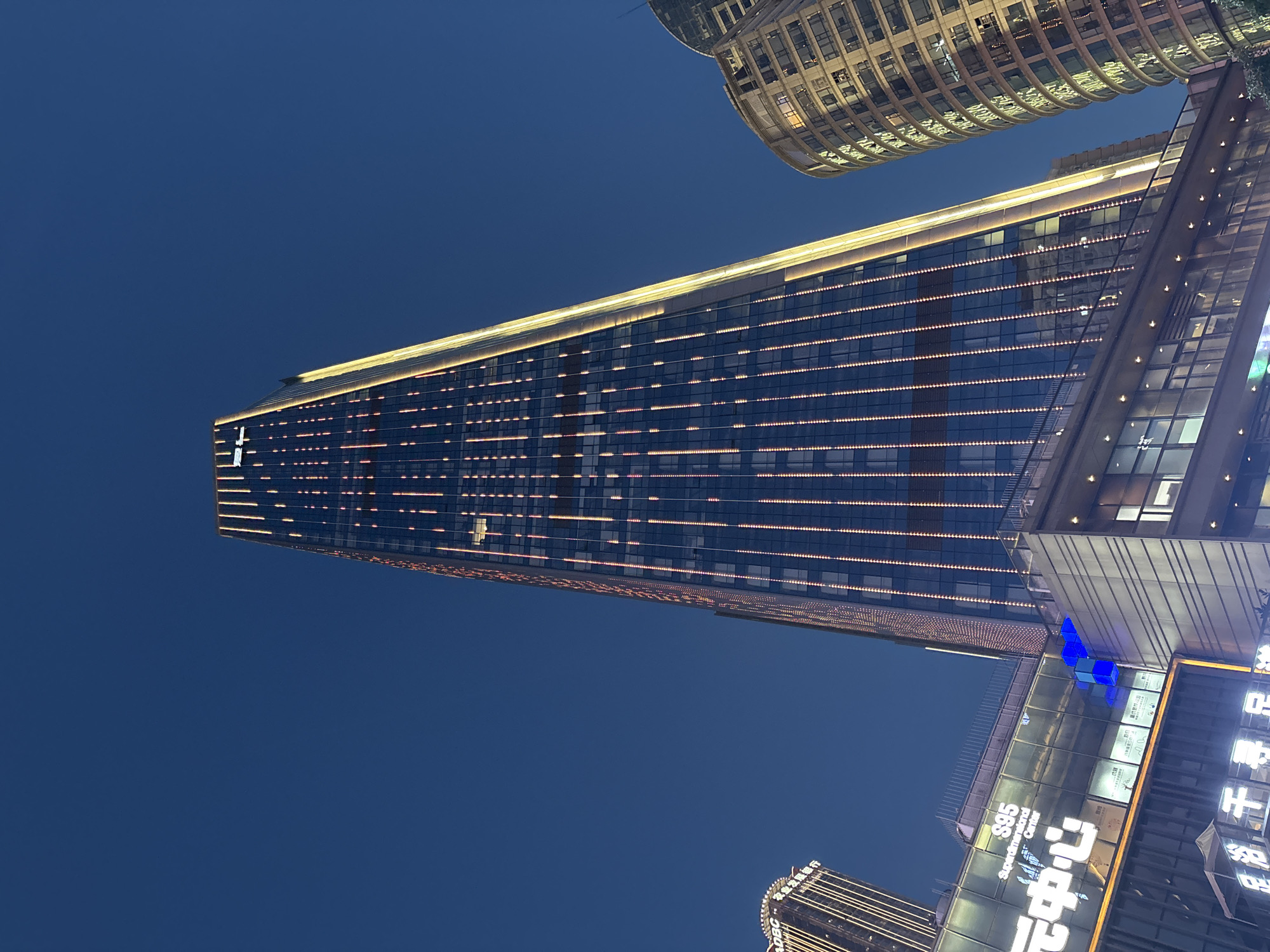The image shows a tall building in the Yuzhong District of Chongqing, China at night. The building is rectangular, with horizontal rows of lights illuminating its facade. Another, shorter building, curved and also lit up at night, is located near the first. Several storefronts with visible signage are at the base of the building. The background sky is dark blue.