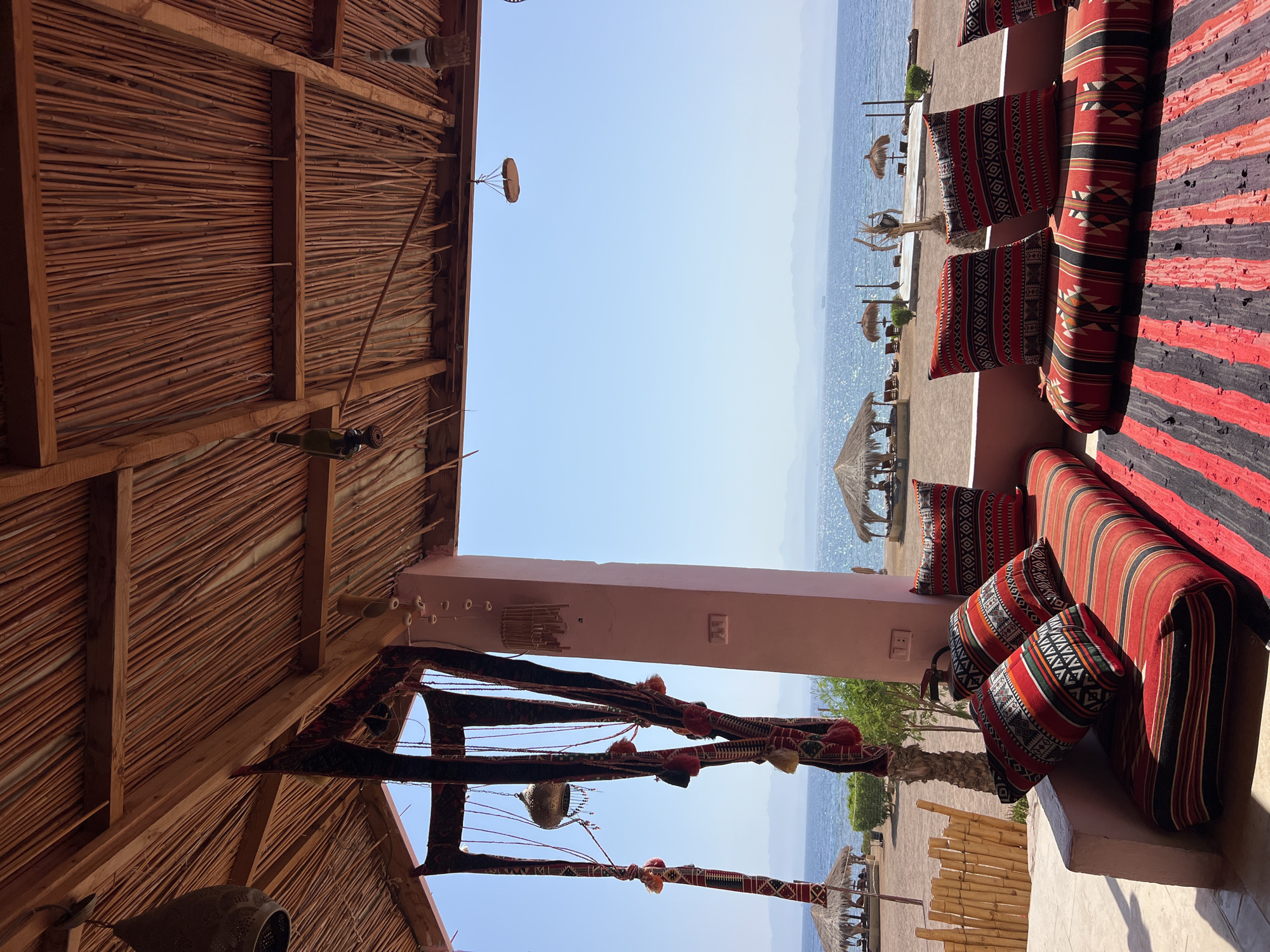 This image depicts an outdoor seating area with a view of the Red Sea, likely in Nueiba, Egypt. The foreground features a comfortable seating arrangement with cushions and a rug, all adorned with traditional geometric patterns in red, black, and white. A thatched roof supported by wooden beams covers the area, with decorative elements hanging from it. Beyond the seating, a sandy beach is visible, dotted with straw-roofed huts and lounge chairs, indicating a resort or a similar tourist-oriented establishment. The calm, blue waters of the Red Sea stretch out to the horizon, where hazy mountains are visible. The sky is a clear, bright blue. The overall impression is one of a tranquil, seaside relaxation spot.