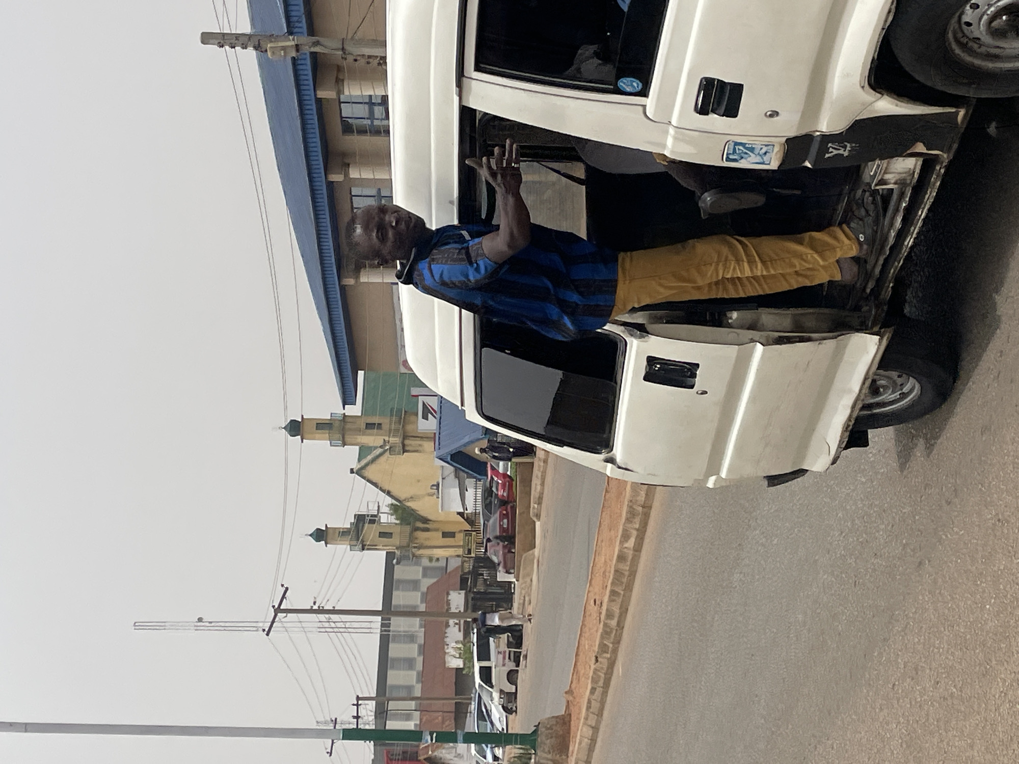 The image shows a man standing by a white van, seemingly waving or gesturing with his right hand. The man is wearing a blue and black striped shirt and yellow pants. He is positioned by the open driver's side door of the van. The van itself appears to be old and possibly used for commercial transport. In the background, there is a street scene with buildings, power lines, and other vehicles. There is a mosque in the background. The image was taken in the city of Jos, Nigeria.
