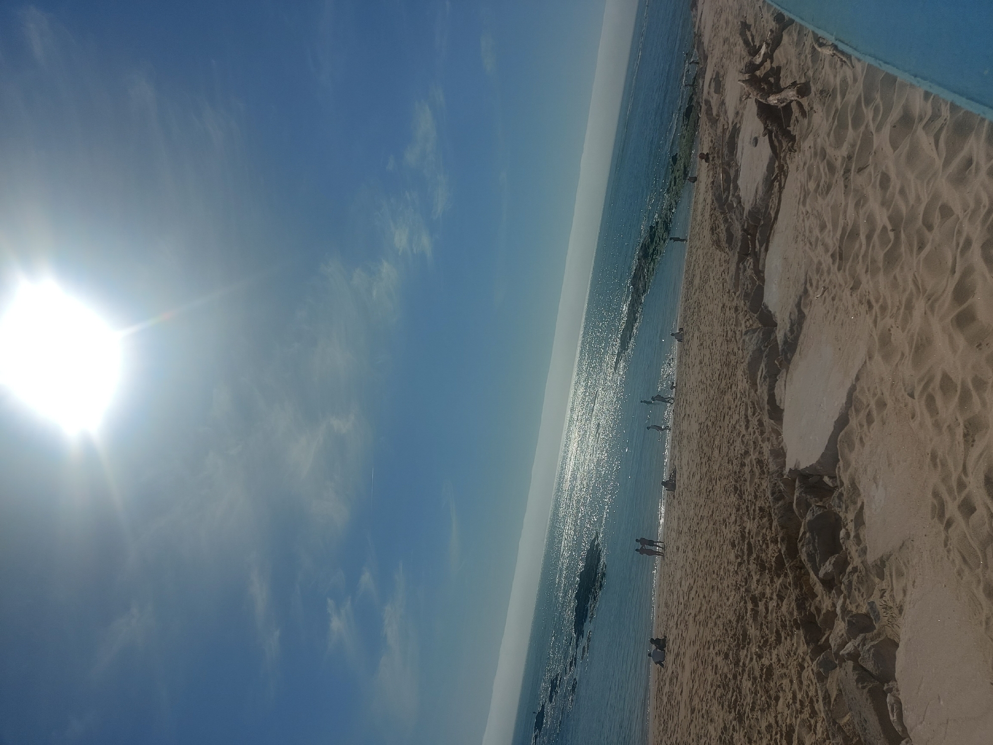 The image captures a beach scene in Figueira da Foz, Portugal, under a bright, sunny sky with scattered clouds. The sun, positioned to the left of the frame, casts a strong glare. The Atlantic Ocean stretches across the central part of the image, reflecting sunlight. Along the right side, there is a sandy beach. Several individuals are visible on the beach and in the water, some standing or walking along the shore, others appearing to be wading or swimming. The environment is characterized by sand, rocks, and the ocean meeting the shoreline. There are some rock formations on the beach. Some driftwood is at the upper right corner of the image. A blue structure is visible at the upper right corner of the frame. The scene depicts a typical sunny day at the beach.